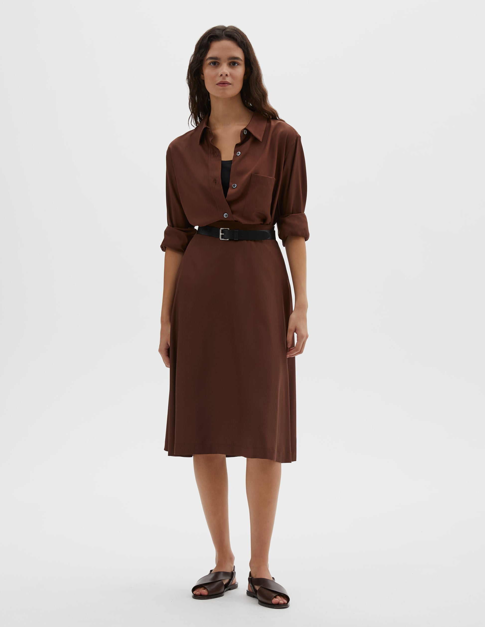 Full body image, front view of model wearing brown matt silk skirt, with side closure and horn button detailing on the waist. Styled with matching brown shirt, black cotton vest, black leather belt and brown leather sandals.