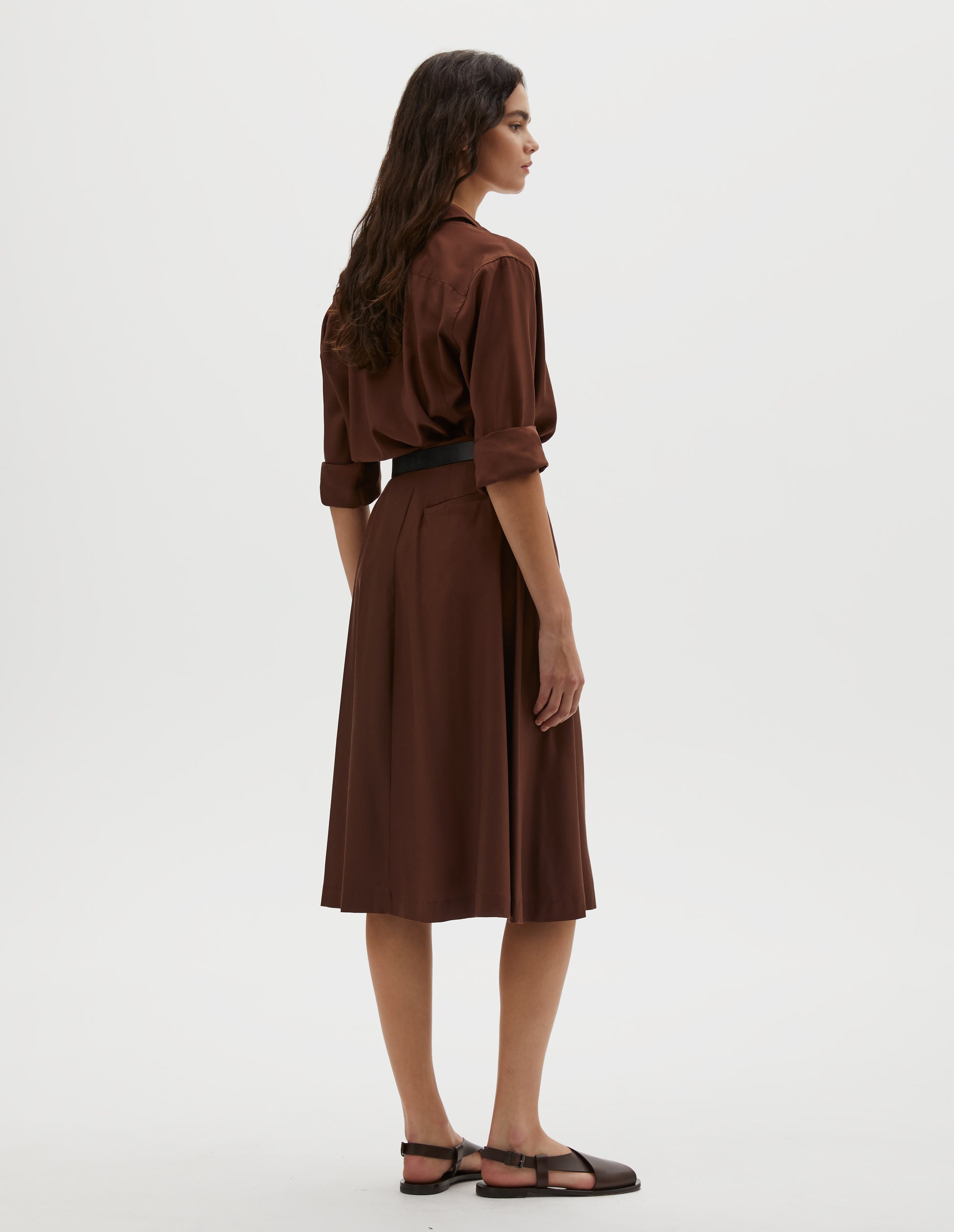 Full body image, back view of model wearing brown matt silk skirt. Styled with matching brown shirt, black leather belt and brown leather sandals.