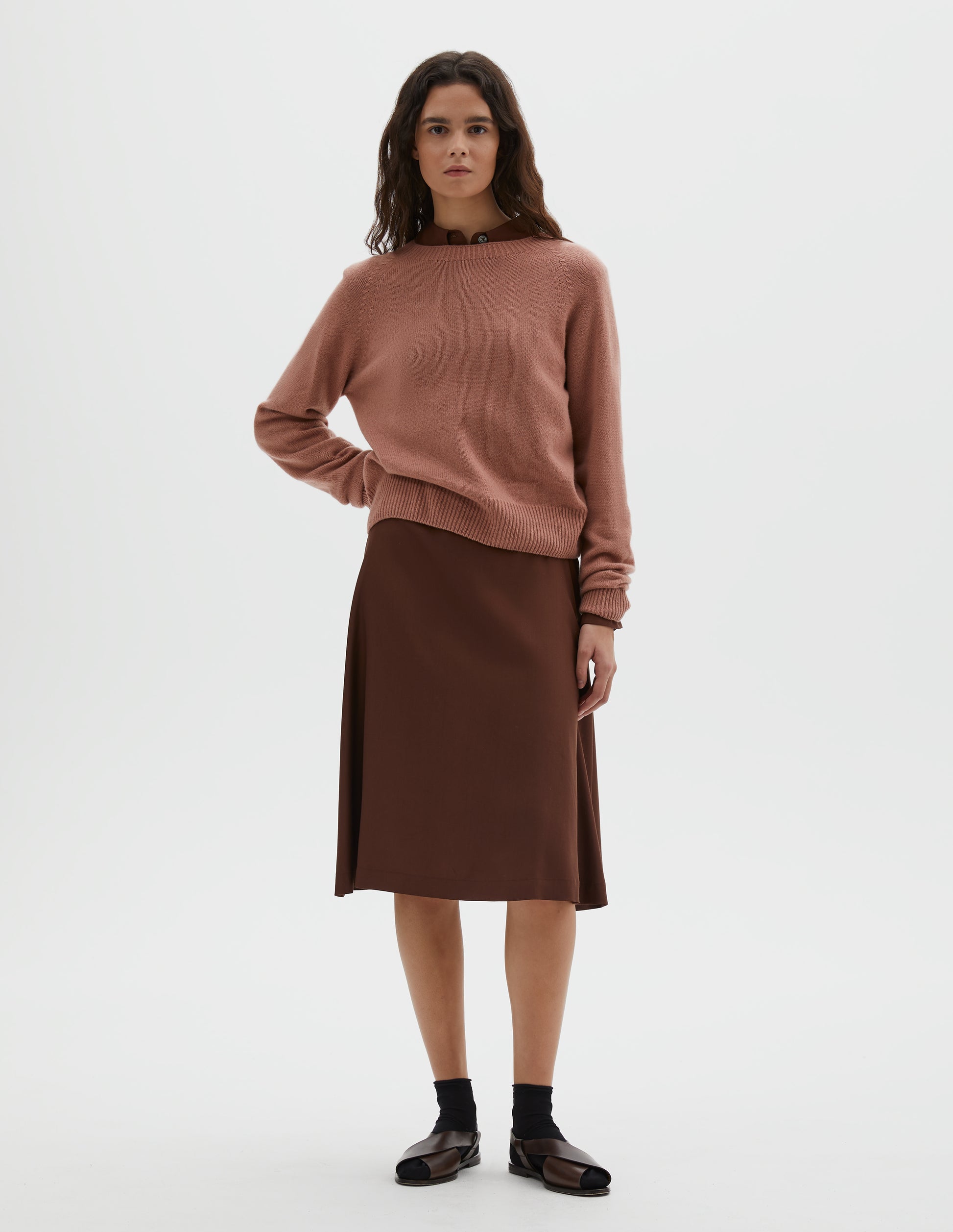 Full body image, front view of model wearing pink long sleeve merino cashmere jumper, with raglan sleeve and contrasting rib on neck, cuffs and hem. Styled over a brown shirt and matching brown skirt, with black socks and brown leather sandals.