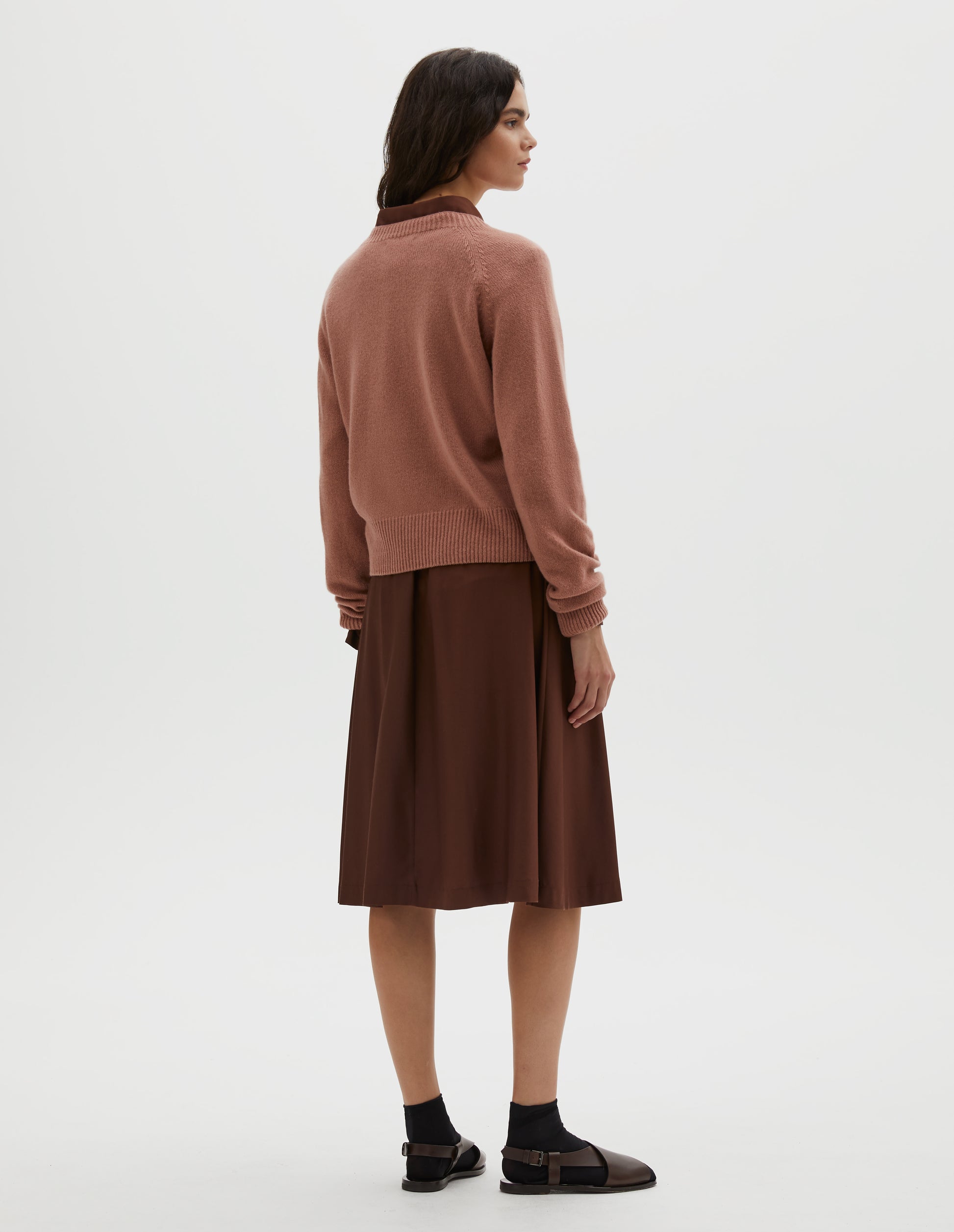 Full body image, back view of model wearing pink long sleeve merino cashmere jumper, with raglan sleeve and contrasting rib on neck, cuffs and hem. Styled over a brown shirt and matching brown skirt, with black socks and brown leather sandals.