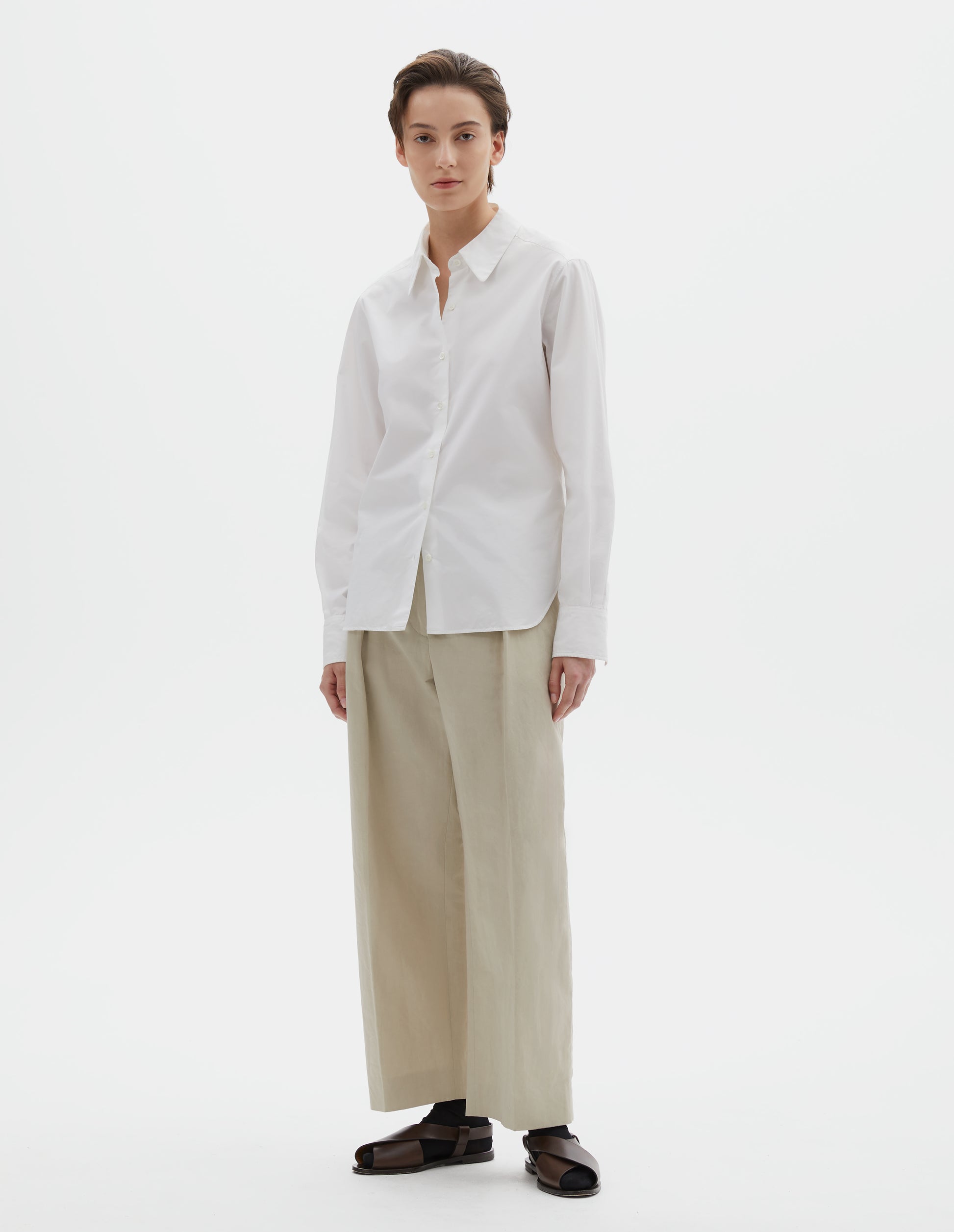 Full body image, front view of model wearing white shirt with raised placket and mother of pearl buttons, patch pocket on chest. Styled with beige pleated trousers, black socks and brown leather sandals.