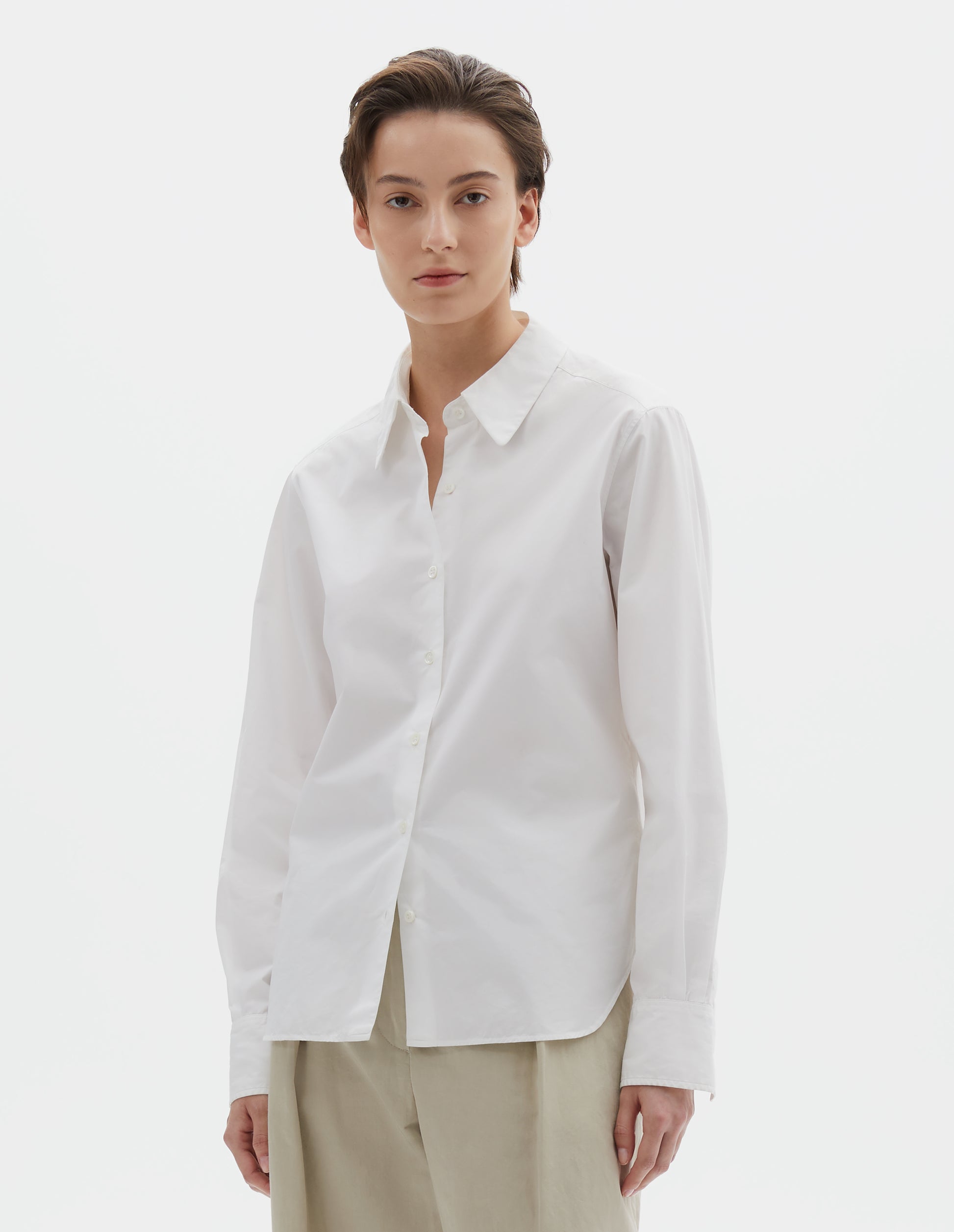Cropped image, front view of model wearing white shirt with raised placket and mother of pearl buttons, patch pocket on chest. Styled with beige pleated trousers.