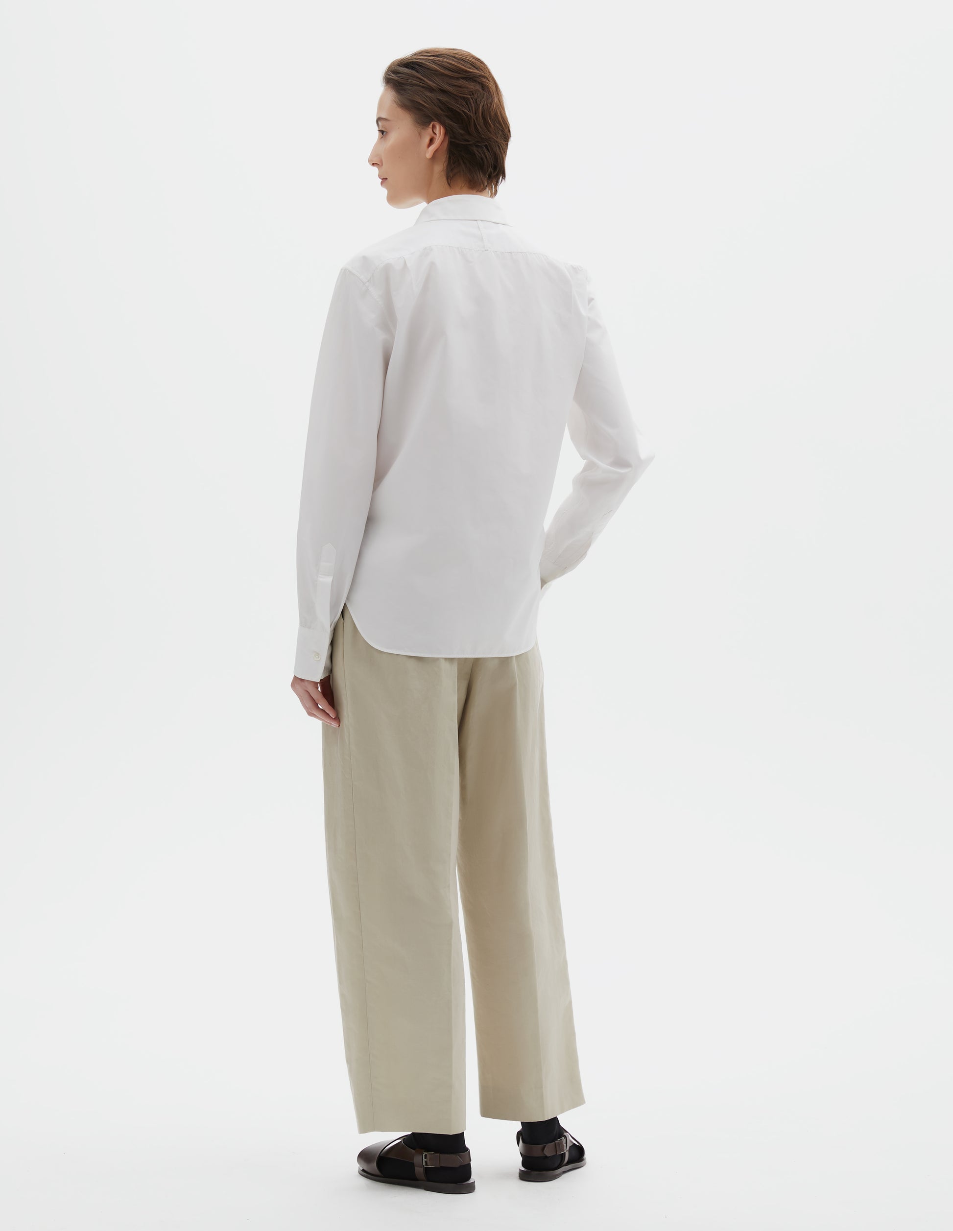 Full body image, back view of model wearing white shirt with mother of pearl buttons on cuffs. Styled beige pleated trousers, black socks and brown leather sandals.
