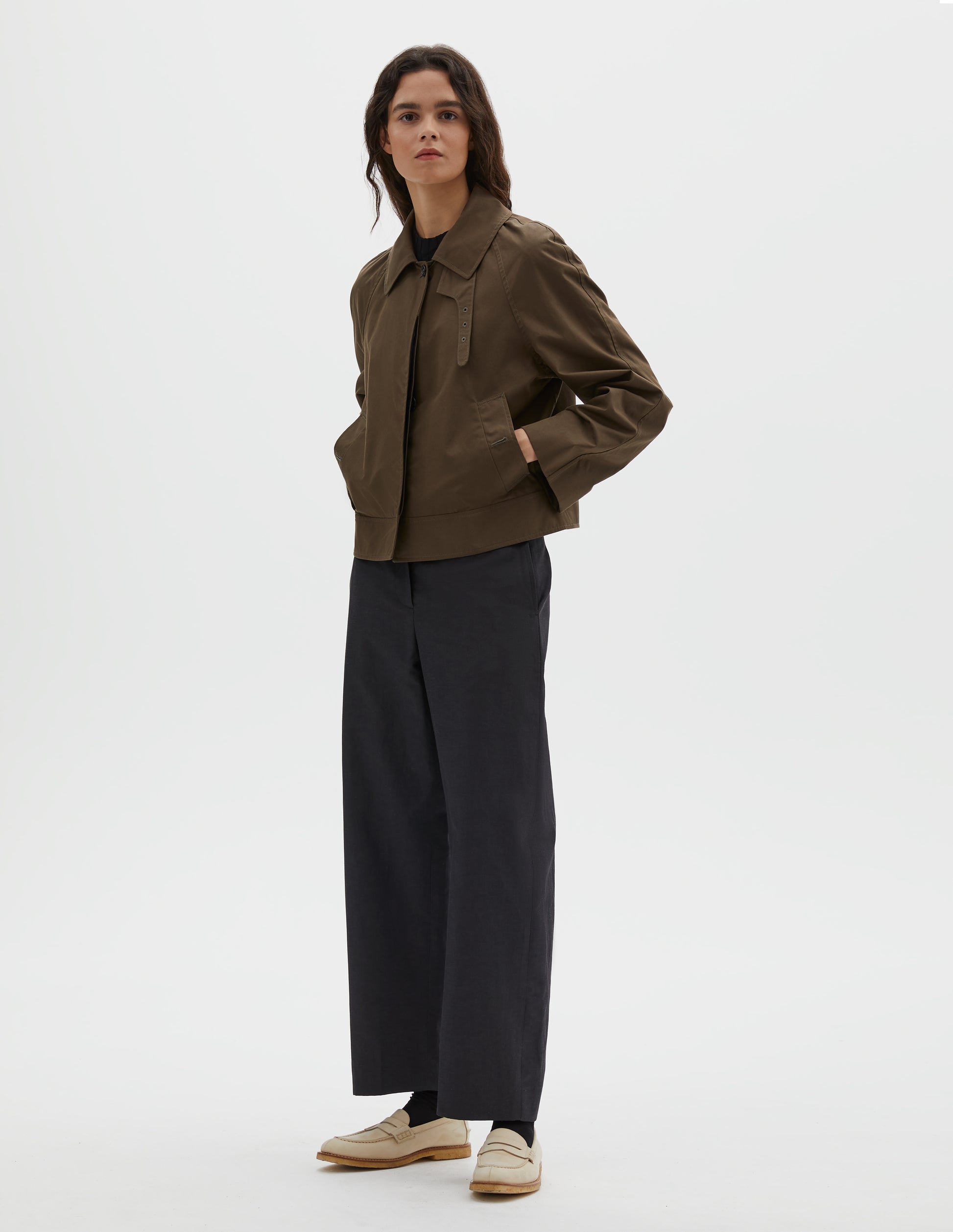 Full body image, front view of model wearing greenish brown ventile cotton poplin long sleeve cropped trench, with horn button closure, slant side pockets and adjustable storm tab. Styled over black t-shirt, with dark navy trousers, black socks and beige loafers.