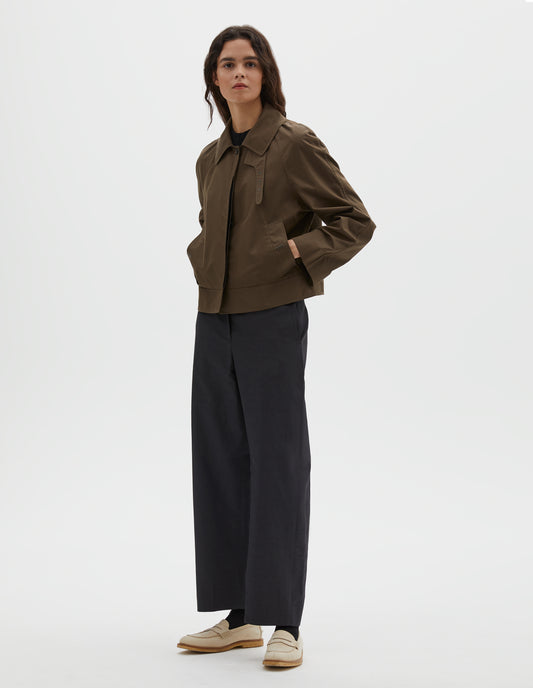 Full body image, front view of model wearing greenish brown ventile cotton poplin long sleeve cropped trench, with horn button closure, slant side pockets and adjustable storm tab. Styled over black t-shirt, with dark navy trousers, black socks and beige loafers.