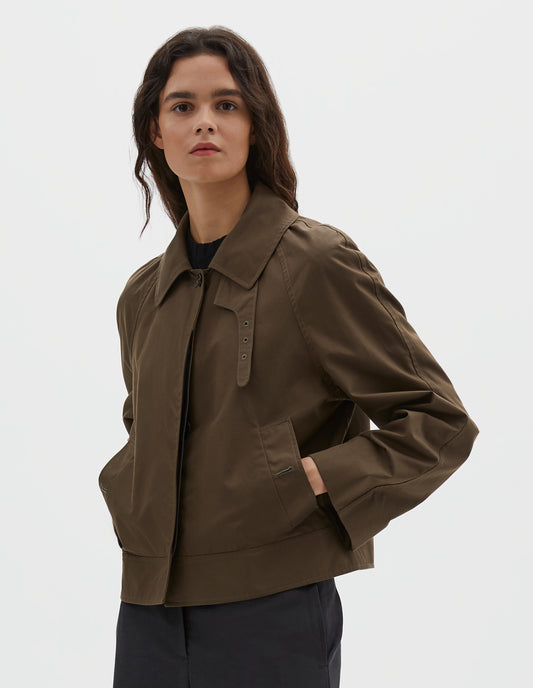 Cropped body image, front view of model wearing greenish brown ventile cotton poplin long sleeve cropped trench, with horn button closure, slant side pockets and adjustable storm tab. Styled over black t-shirt, with dark navy trousers.