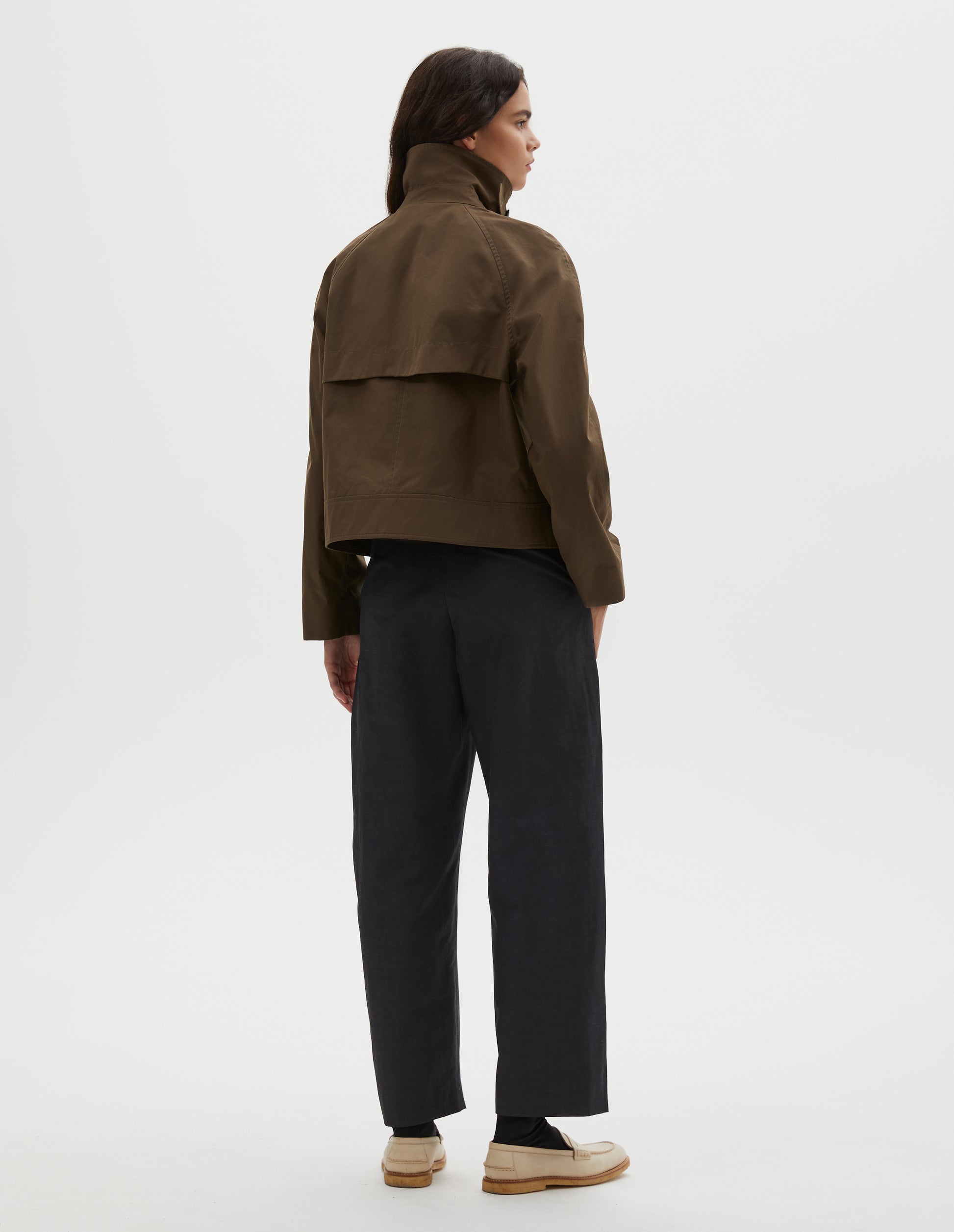 Full body image, back view of model wearing greenish brown ventile cotton poplin long sleeve cropped trench, with storm vent. Styled with dark navy trousers, black socks and beige loafers.