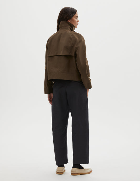 Full body image, back view of model wearing greenish brown ventile cotton poplin long sleeve cropped trench, with storm vent. Styled with dark navy trousers, black socks and beige loafers.