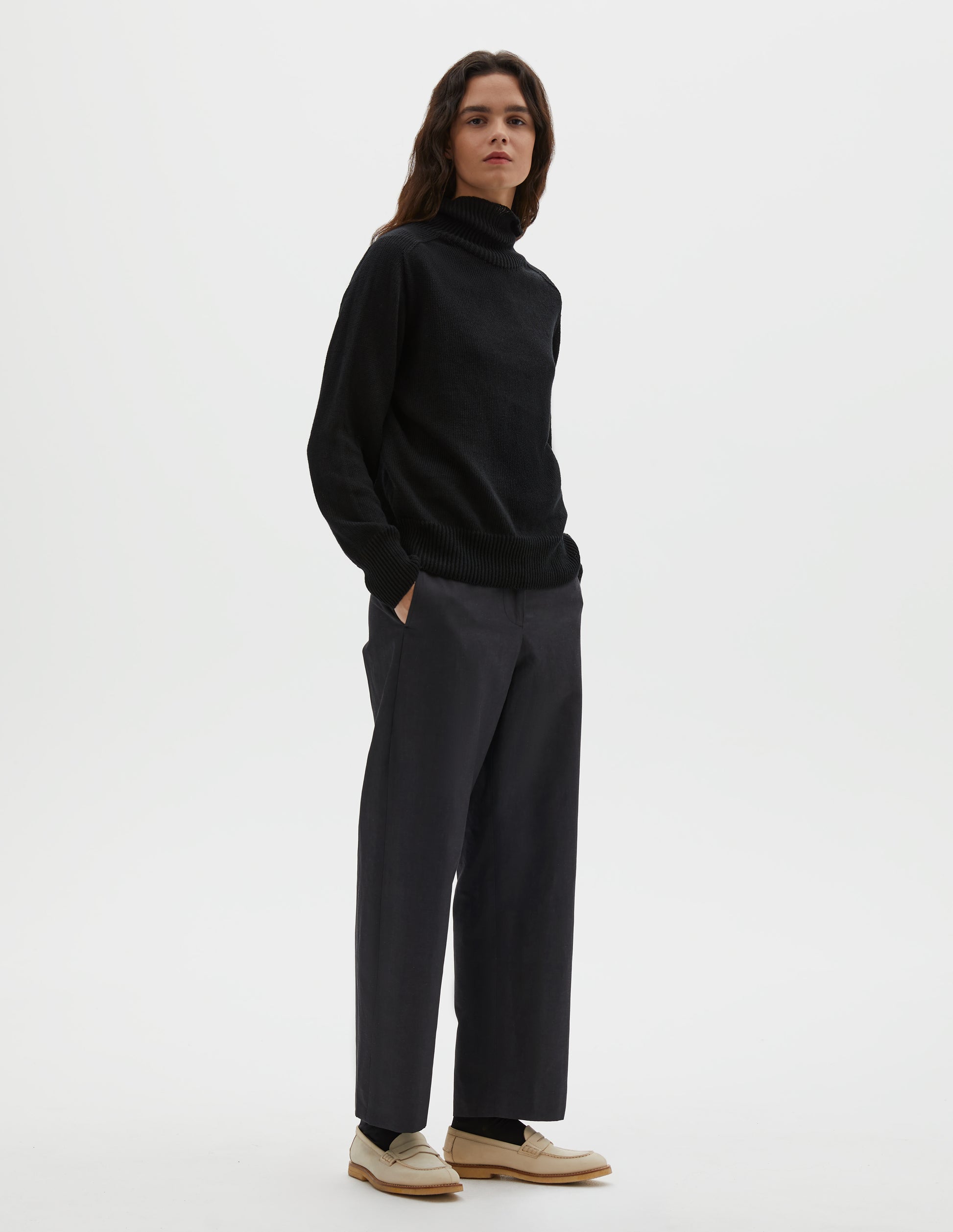 Full body image, front view of model wearing black linen cotton roll neck jumper with long saddle sleeves and contrast rib on neck, cuffs and hem. Styled with black trousers, black socks and beige suede loafers.