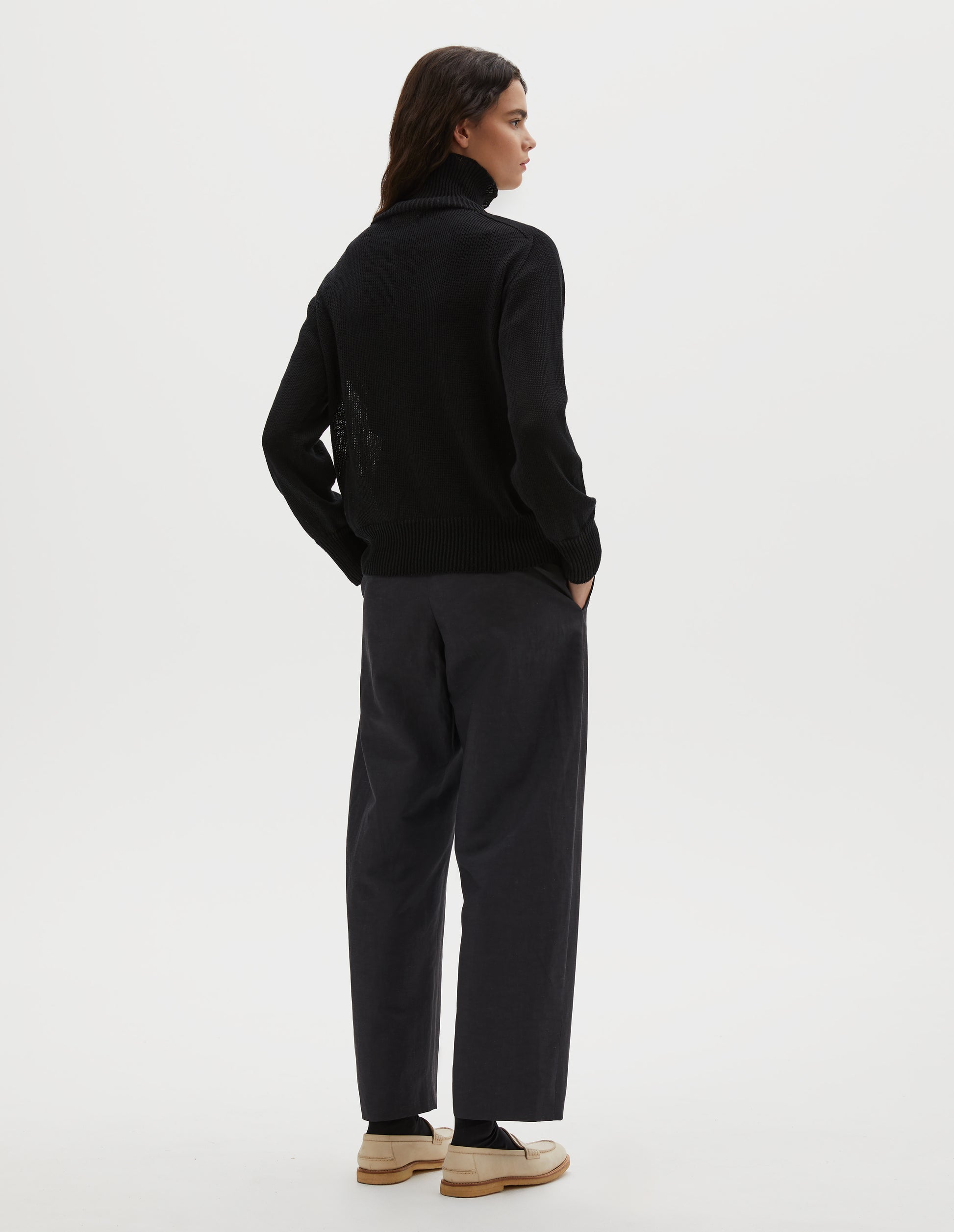 Full body image, back view of model wearing black linen cotton roll neck jumper with long saddle sleeves and contrast rib on neck, cuffs and hem. Styled with black trousers, black socks and beige suede loafers.