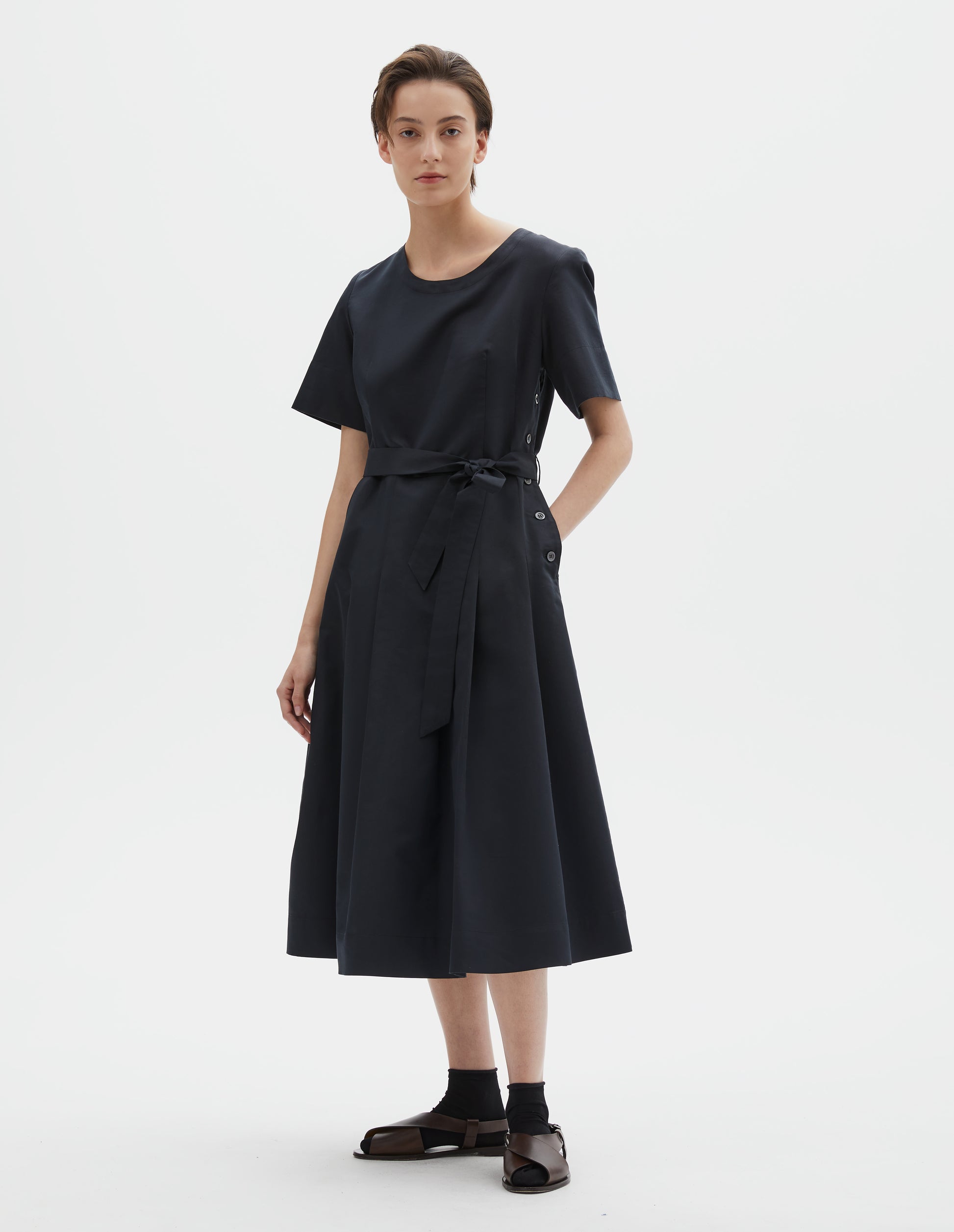 Full body image, front view of model wearing black short sleeve a-line cotton silk satin dress, with fitted waist, pleated skirt, detachable matching belt and mother of pearl button fastening down one side. Styled with black socks and brown leather sandals.