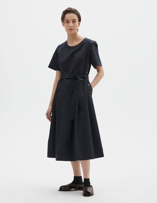 Full body image, front view of model wearing black short sleeve a-line cotton silk satin dress, with fitted waist, pleated skirt, detachable matching belt and mother of pearl button fastening down one side. Styled with black socks and brown leather sandals.