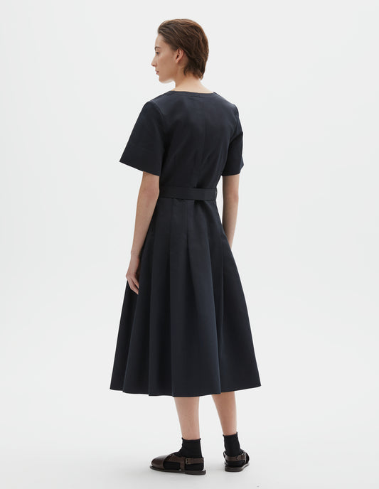Full body image, back view of model wearing black short sleeve a-line cotton silk satin dress, with fitted waist, pleated skirt, and detachable matching belt. Styled with black socks and brown leather sandals.