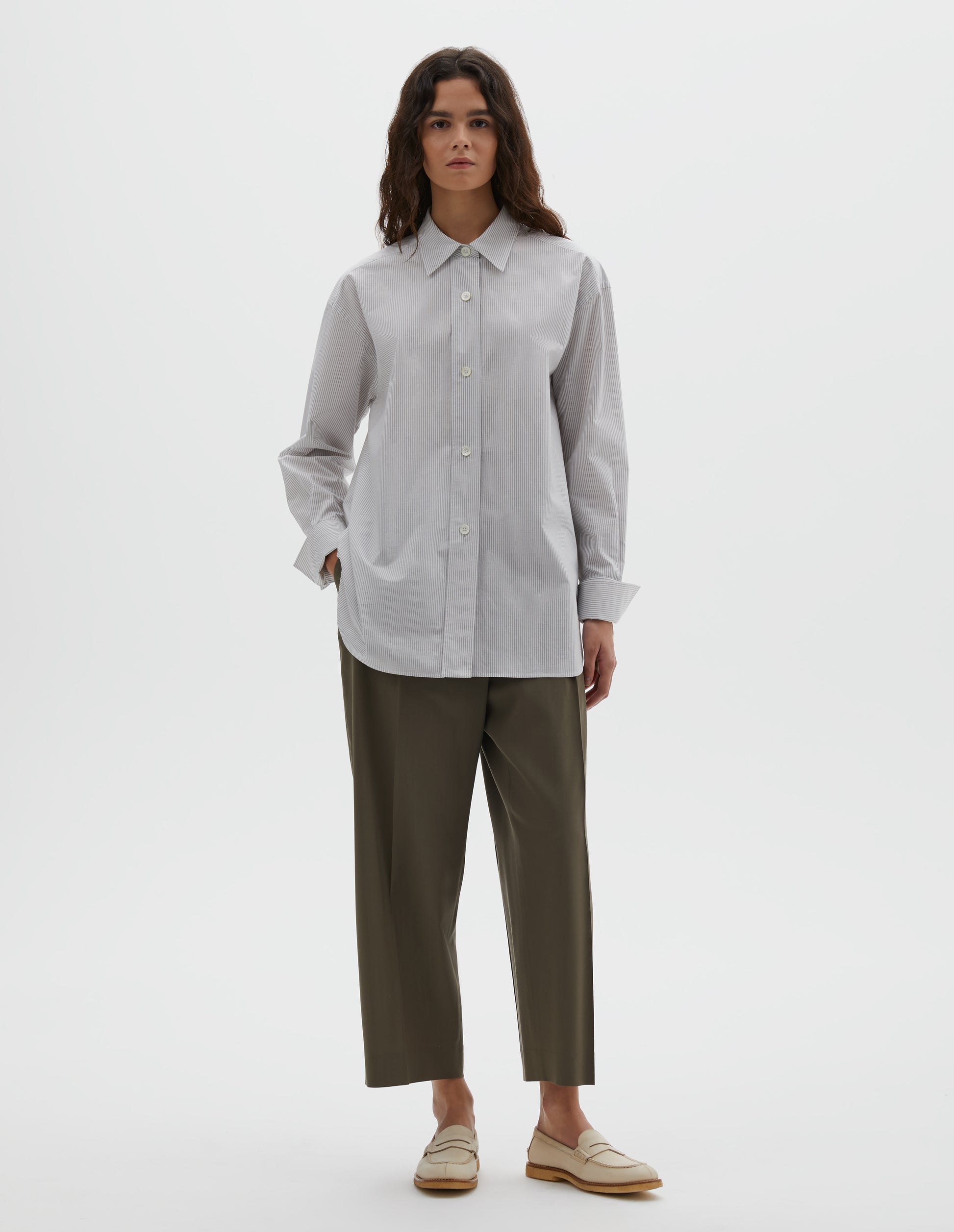 Full body image, front view of model wearing white and grey stripe relaxed cut and dropped shoulder cotton shirt, with collar, wide, raised placket and button fastening turned back double cuffs. Styled with brown trousers and beige shoes.