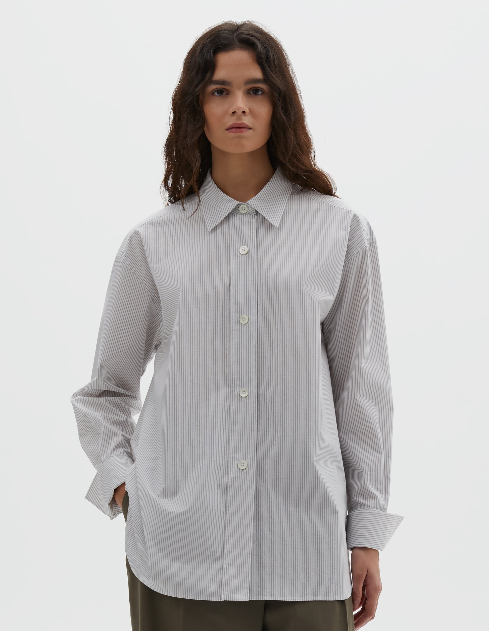 Cropped body image, front view of model wearing white and grey stripe relaxed cut and dropped shoulder cotton shirt, with collar, wide, raised placket and button fastening turned back double cuffs. Styled with brown trousers.