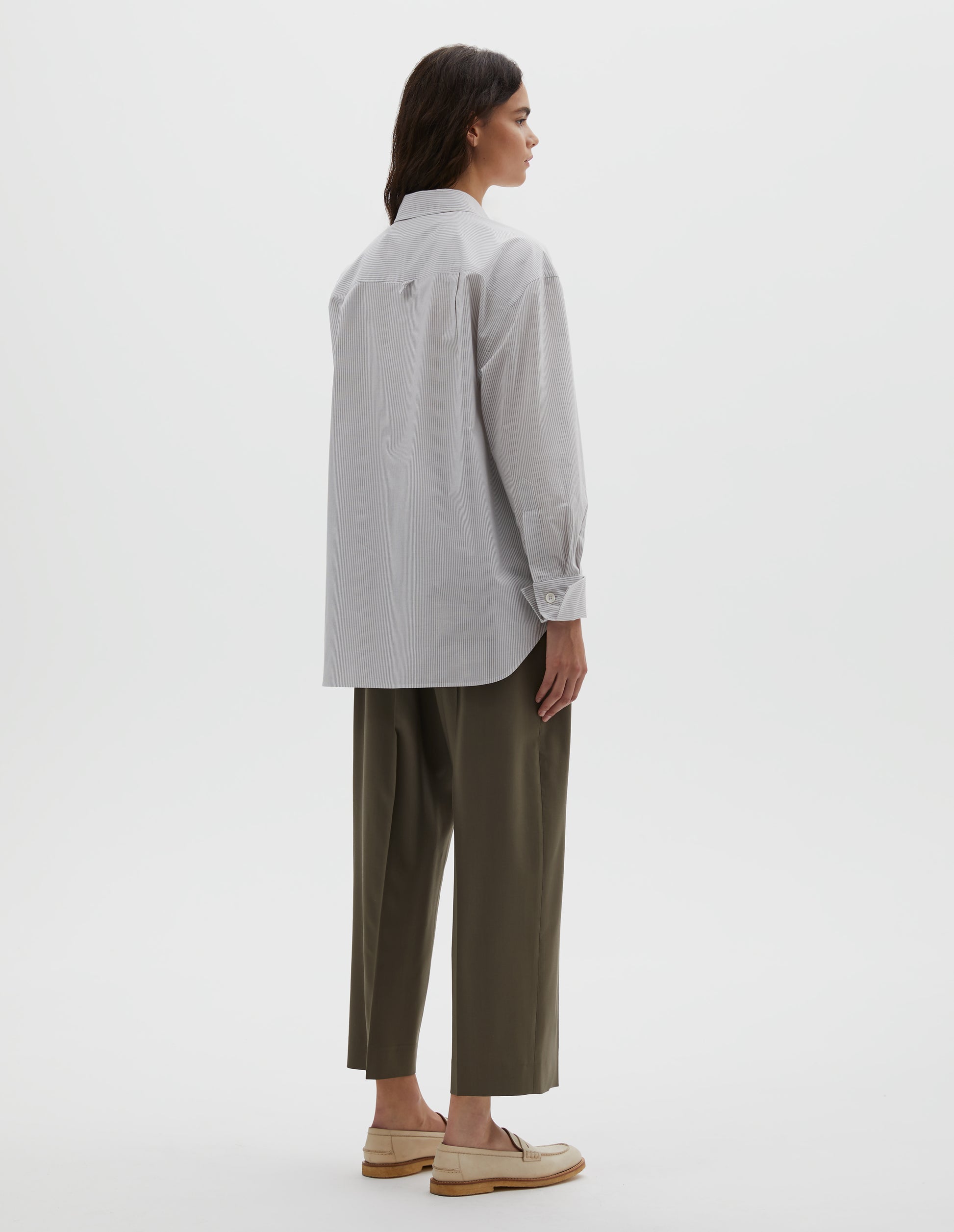 Full body image, back view of model wearing white and grey stripe relaxed cut and dropped shoulder cotton shirt, with collar,  hanger loop detail on the back yoke and button fastening turned back double cuffs. Styled with brown trousers and beige shoes.