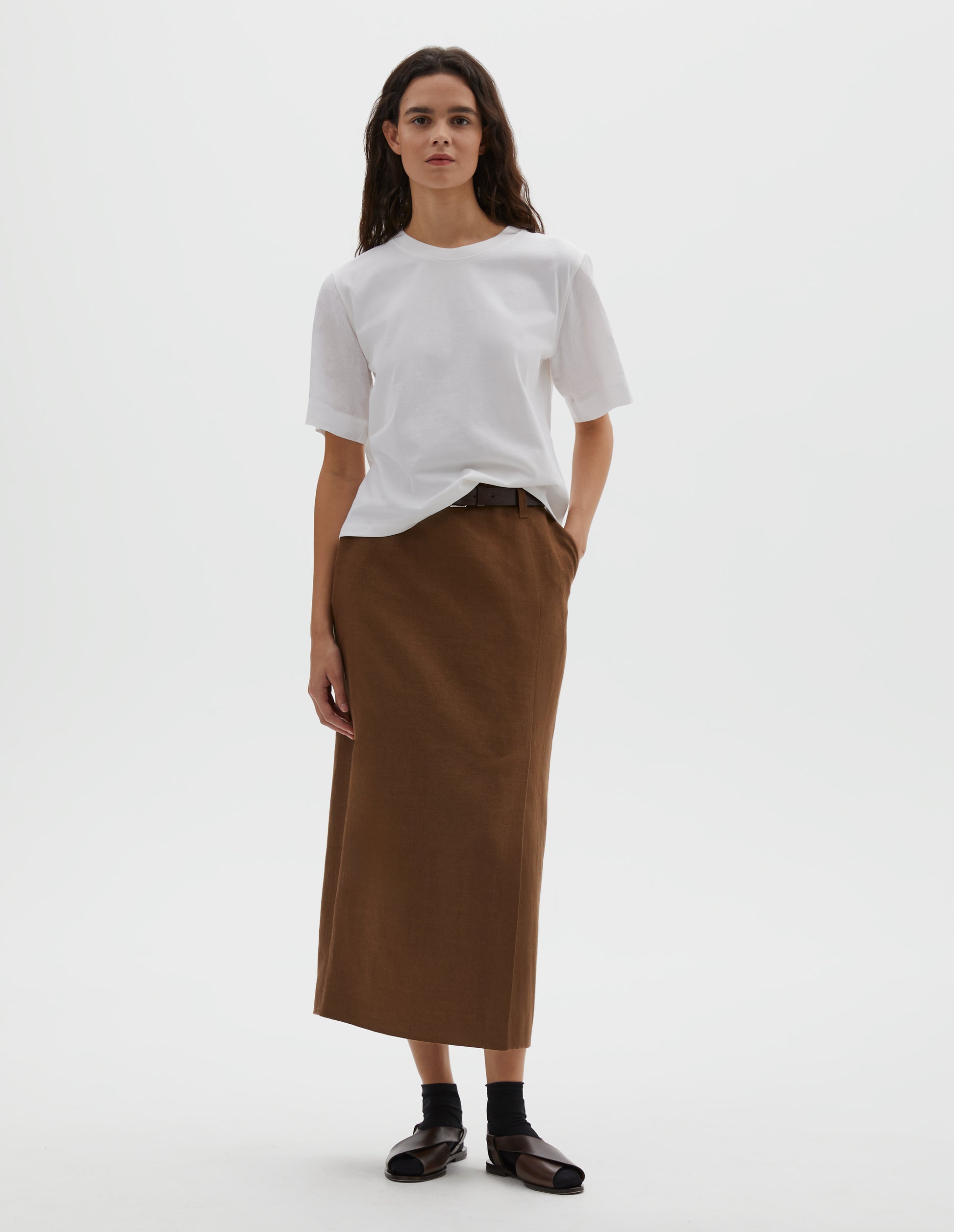 Full body image, front view of model wearing white regular cut t-shirt with contrast Irish shirting linen sleeve. Styled with a brown skirt, black socks and brown sandals.