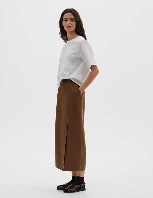 Full body image, front view of model wearing brown pinstripe linen skirt, with offset front vent, concealed side zip fastening, slant side pockets and belt loops. Styled with white cotton t-shirt, black ankle socks and black leather sandals.