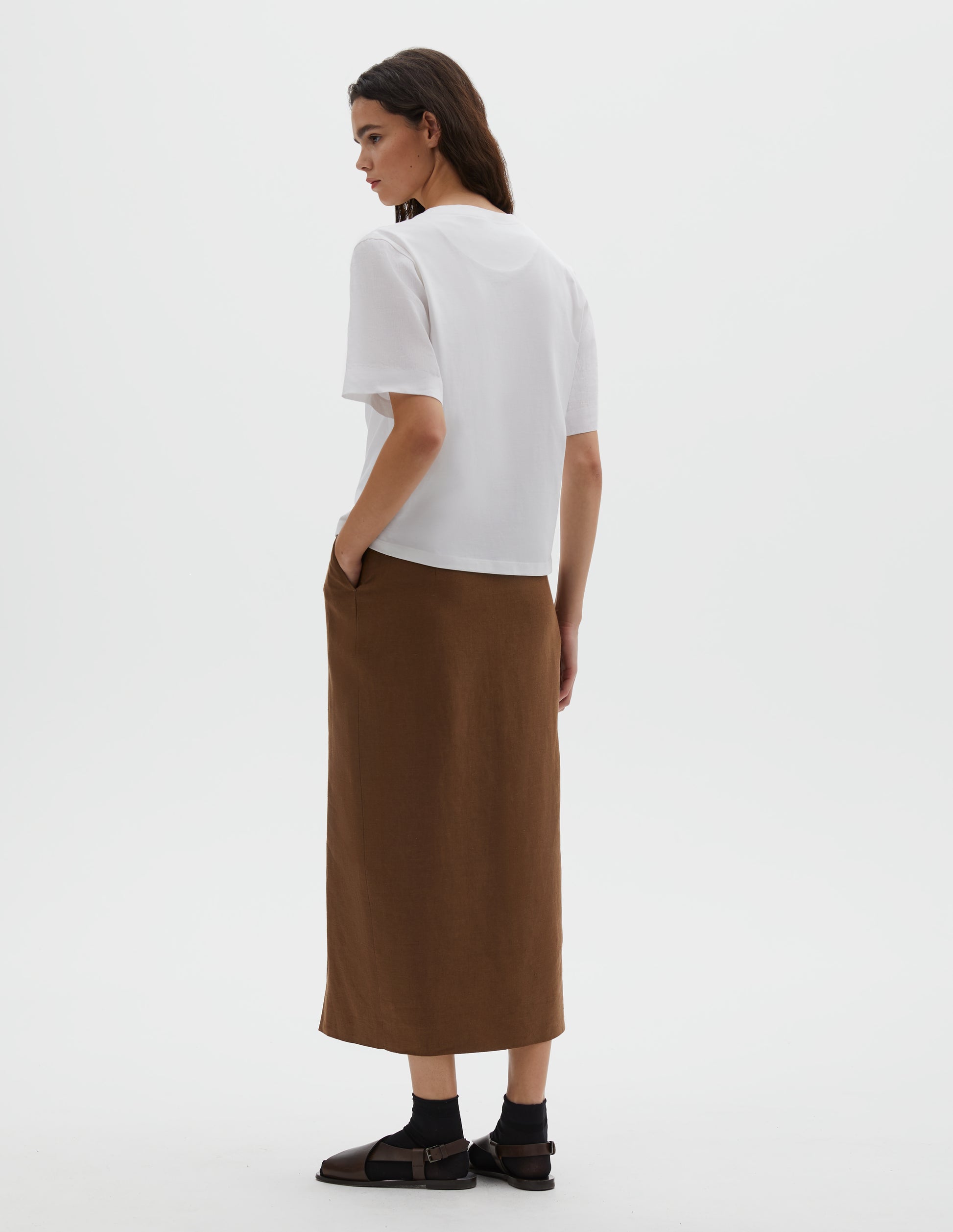 Full body image, back view of model wearing white regular cut t-shirt with contrast Irish shirting linen sleeve. Styled with a brown skirt, black socks and brown sandals.