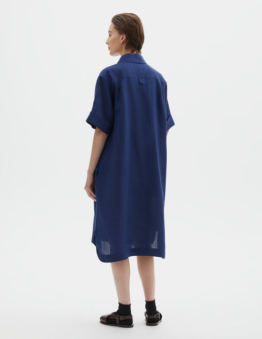 Full body image, back view of model wearing blue linen shirt dress, with concealed side pockets and hanger loop detail on back. Styled with black socks and brown leather sandals.