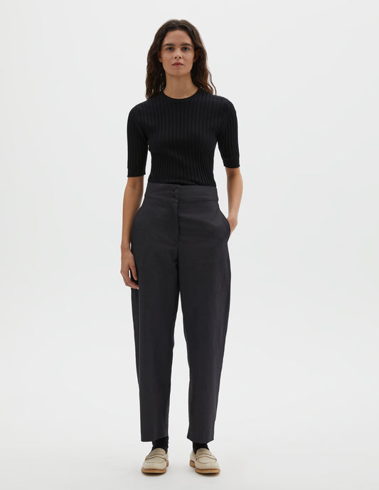 Full body image, front view of model wearing black cotton linen trousers, with deep waistband, double horn button closure and slant side pockets. Styled with black ribbed knitted t-shirt, black socks and beige suede loafers.