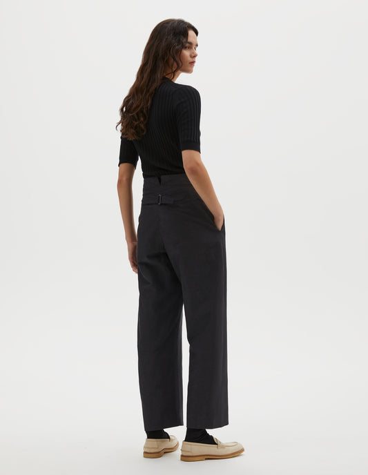 Full body image, back view of model wearing black cotton linen trousers, with deep waistband and cinch adjustor on centre back seam. Styled with black ribbed knitted t-shirt, black socks and beige suede loafers.