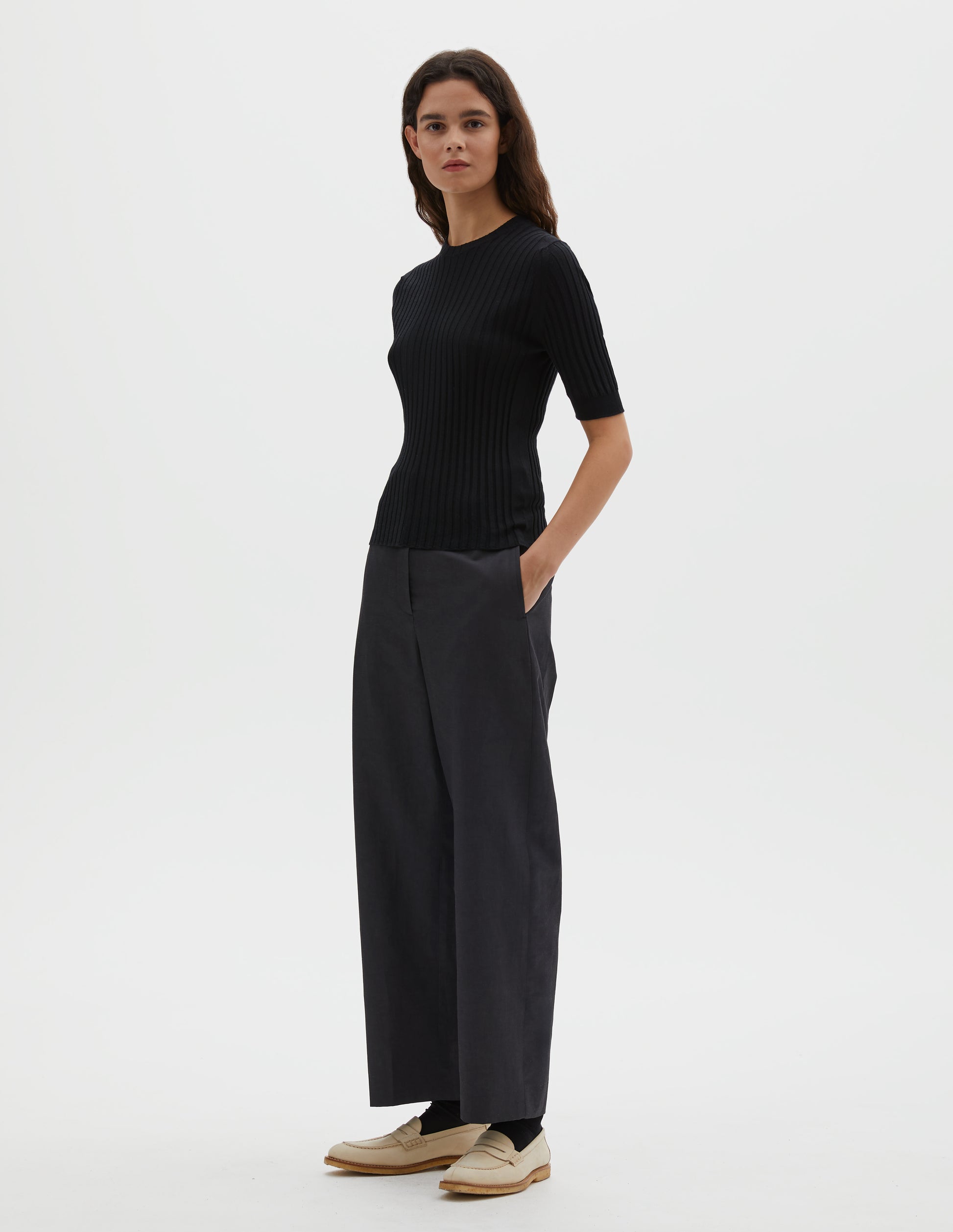 Full body image, front view of model wearing black knitted ribbed t-shirt with round neck and fine rib sleeve cuffs. Styled with grey trousers and white leather loafers.