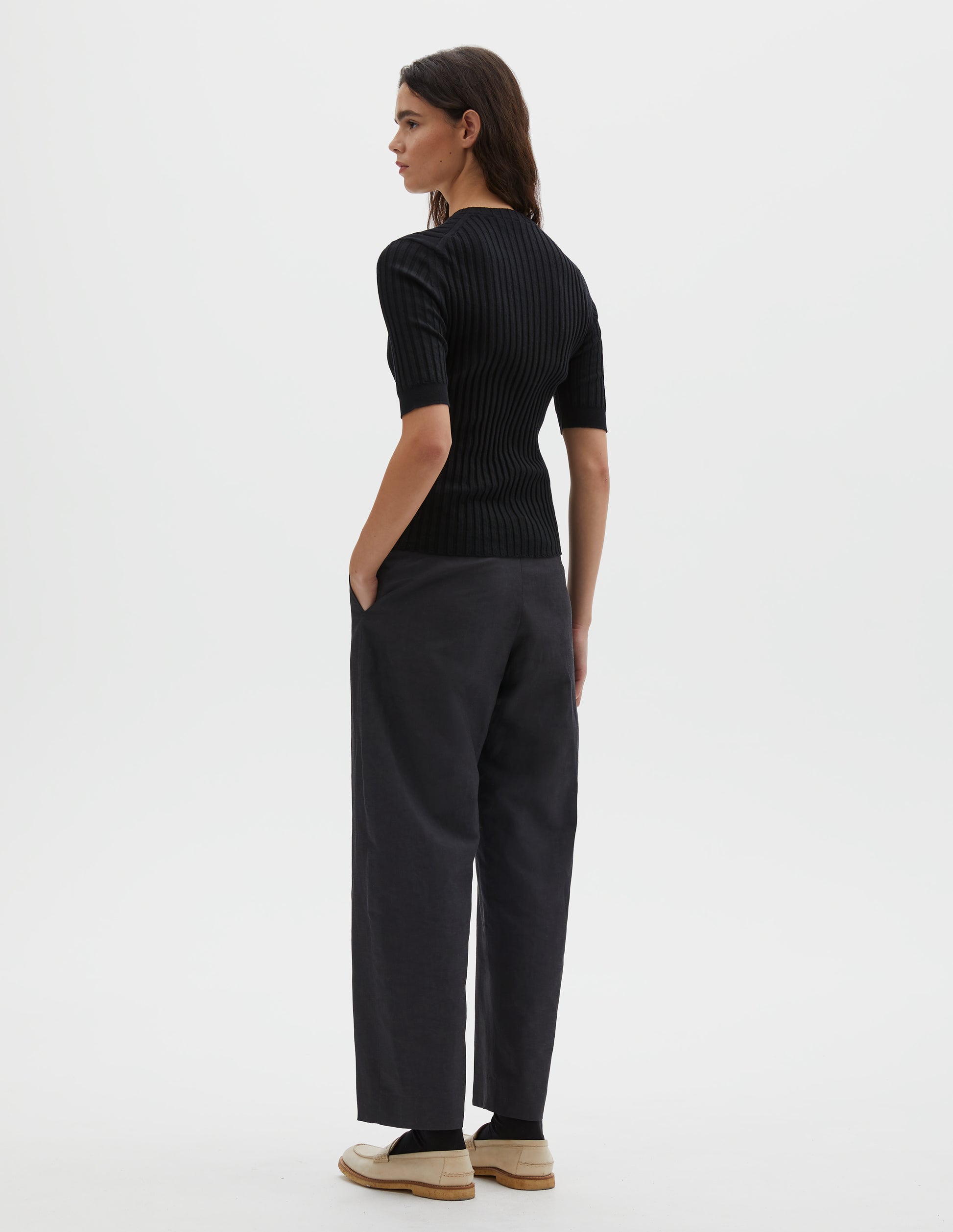 Full body image, back view of model wearing black knitted ribbed t-shirt with round neck and fine rib sleeve cuffs. Styled with grey trousers and white leather loafers.