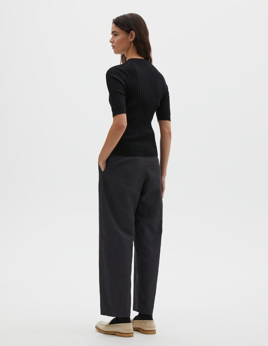 Full body image, back view of model wearing black knitted ribbed t-shirt with round neck and fine rib sleeve cuffs. Styled with grey trousers and white leather loafers.