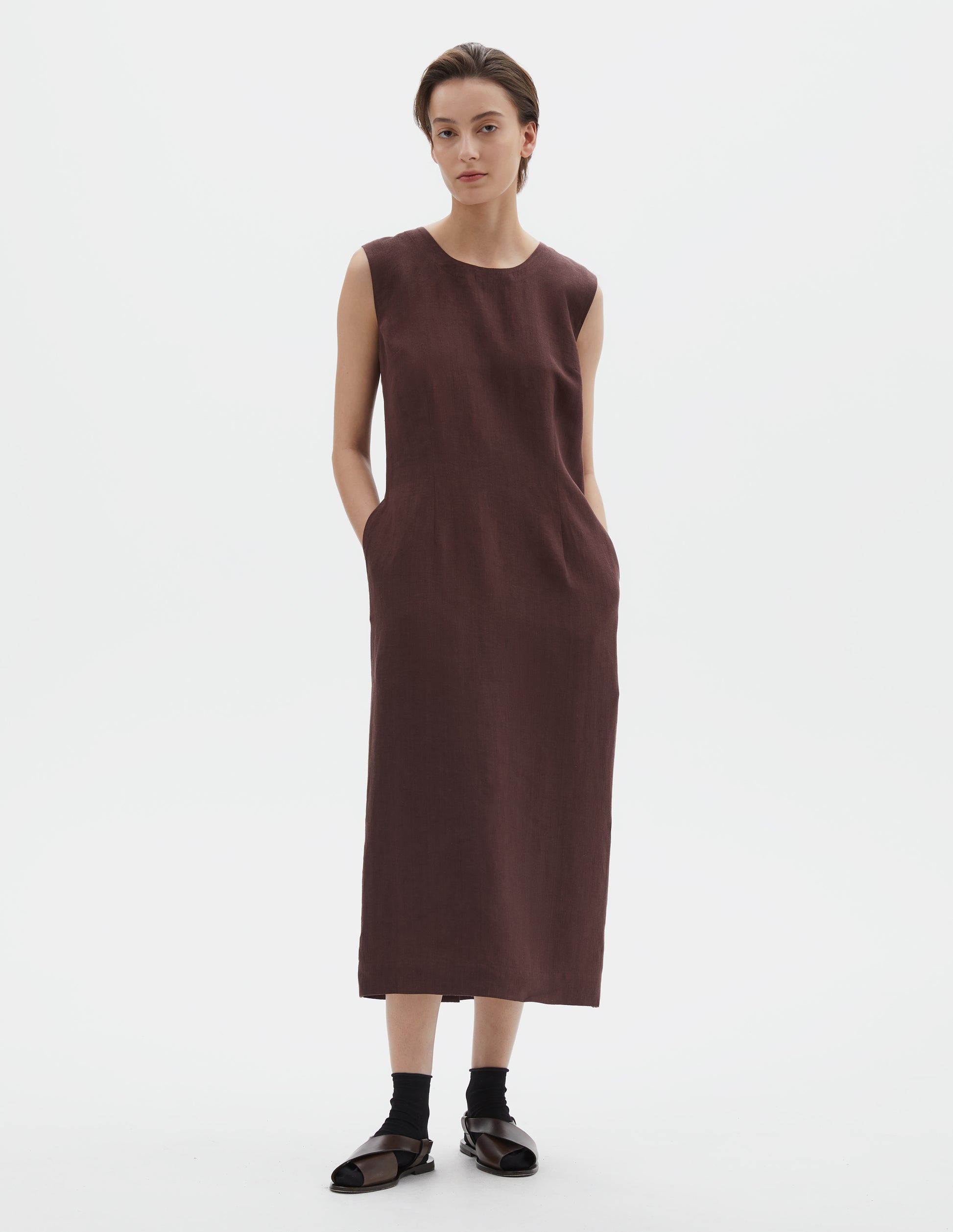 Full body image, front view of model wearing brown regular cut midi length sleeveless dress with side pockets. Styled with black socks and dark brown leather sandals.