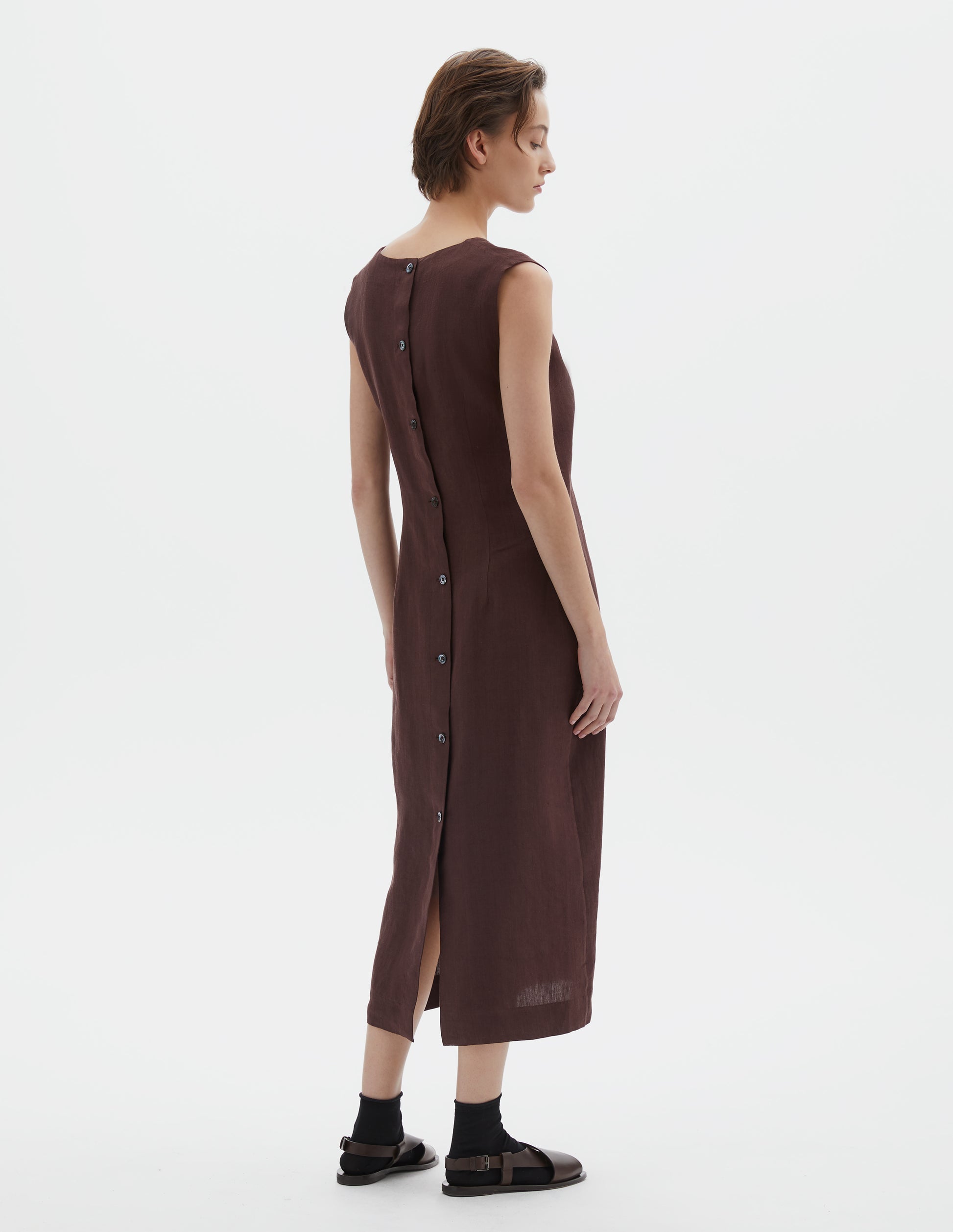 Full body image, back view of model wearing brown regular cut midi length sleeveless dress with mother of pearl buttons on back and deep back vent. Styled with black socks and dark brown leather sandals.
