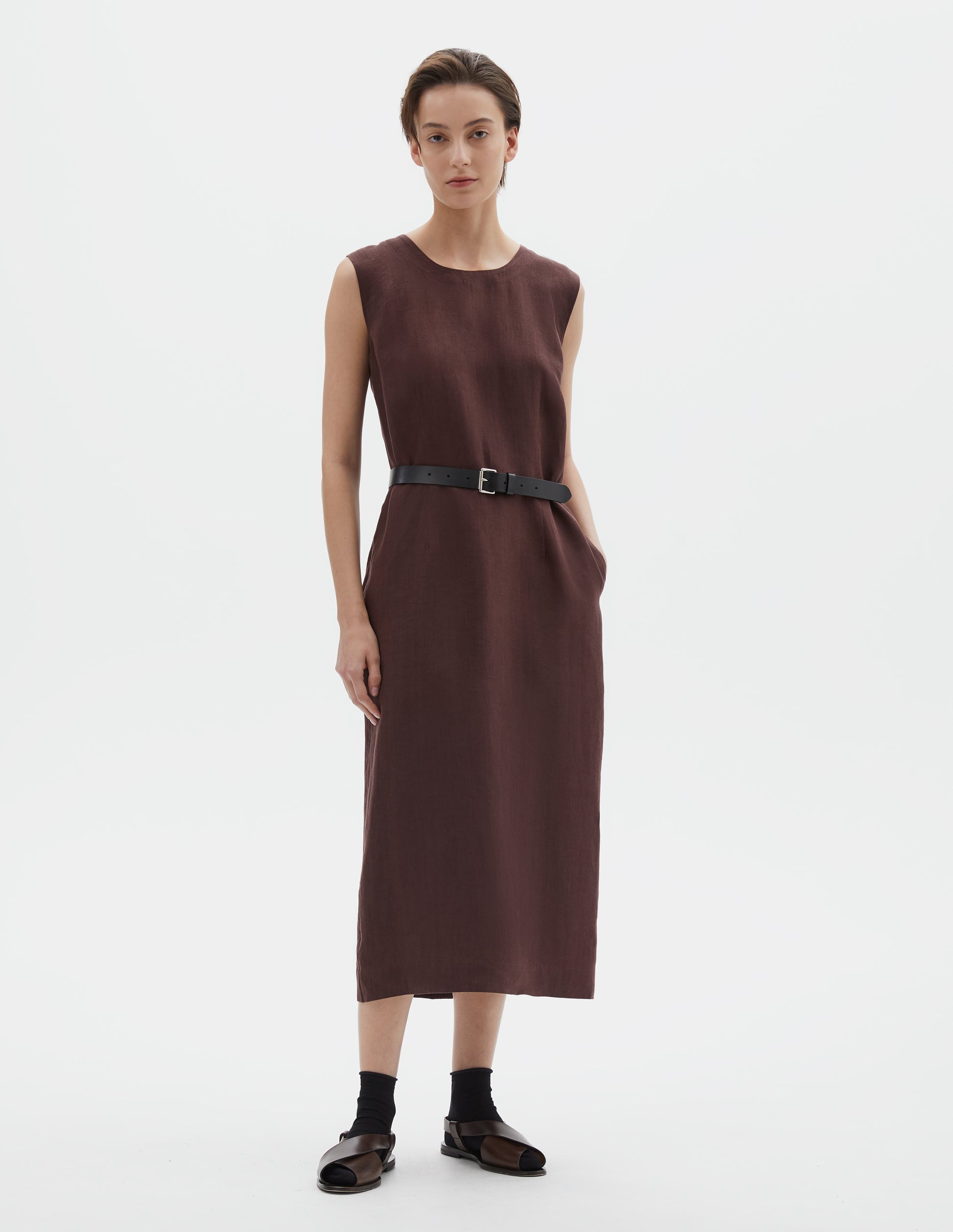 Full body image, front view of model wearing brown regular cut midi length sleeveless dress with side pockets. Styled with black leather belt, black socks and dark brown leather sandals.