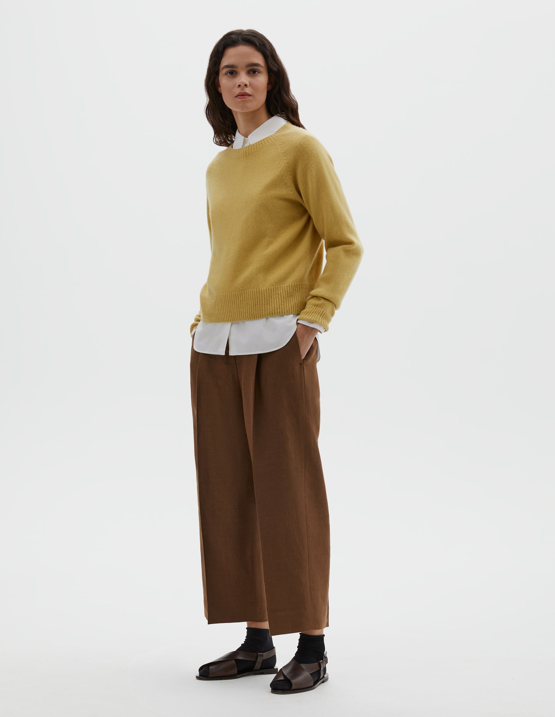 Full body image, front view of model wearing yellow merino cashmere long sleeve jumper, with raglan sleeve and contrast rib on neck, cuffs and hem. Styled over white cotton shirt, with light brown pleated trousers, black ankle socks and brown leather sandals.