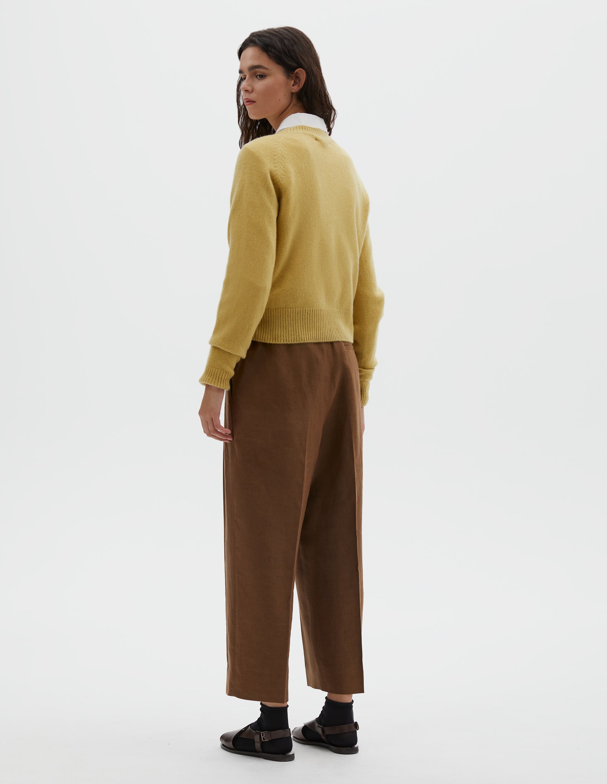 Full body image, back view of model wearing yellow merino cashmere long sleeve jumper, with raglan sleeve and contrast rib on neck, cuffs and hem. Styled over white cotton shirt, with light brown pleated trousers, black ankle socks and brown leather sandals.