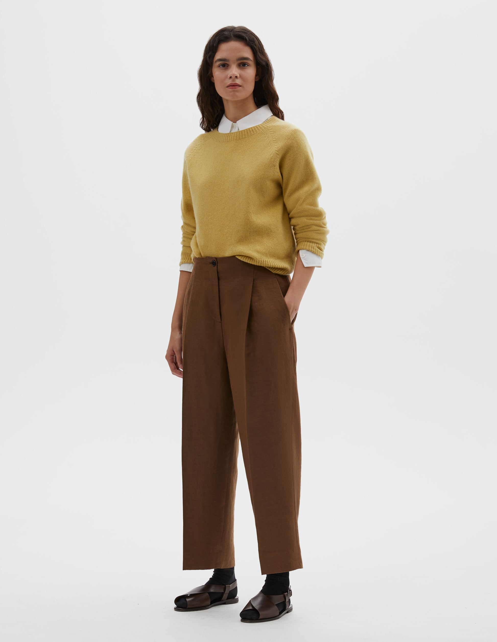 Full body image, front view of model wearing brown linen trousers, with horn button fastening, single pleats and slant side pockets. Styled with  yellow long sleeve jumper, over white cotton shirt, with black ankle socks and brown leather sandals.