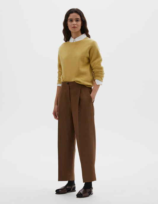 Full body image, front view of model wearing brown linen trousers, with horn button fastening, single pleats and slant side pockets. Styled with  yellow long sleeve jumper, over white cotton shirt, with black ankle socks and brown leather sandals.