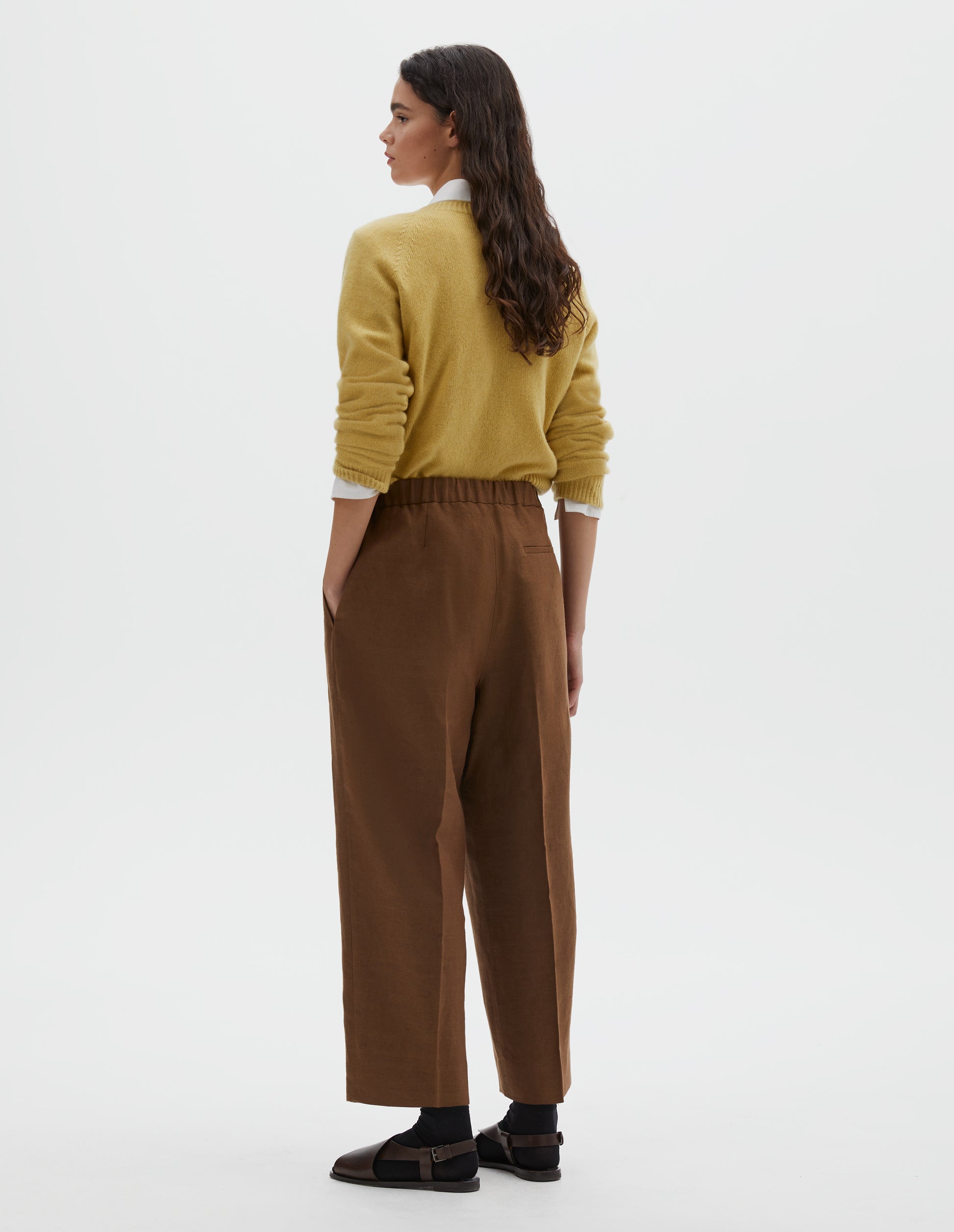 Full body image, back view of model wearing  brown linen trousers, with half elasticated waistband and belt back pocket.  Styled with  yellow long sleeve jumper, over white cotton shirt, with black ankle socks and brown leather sandals.
