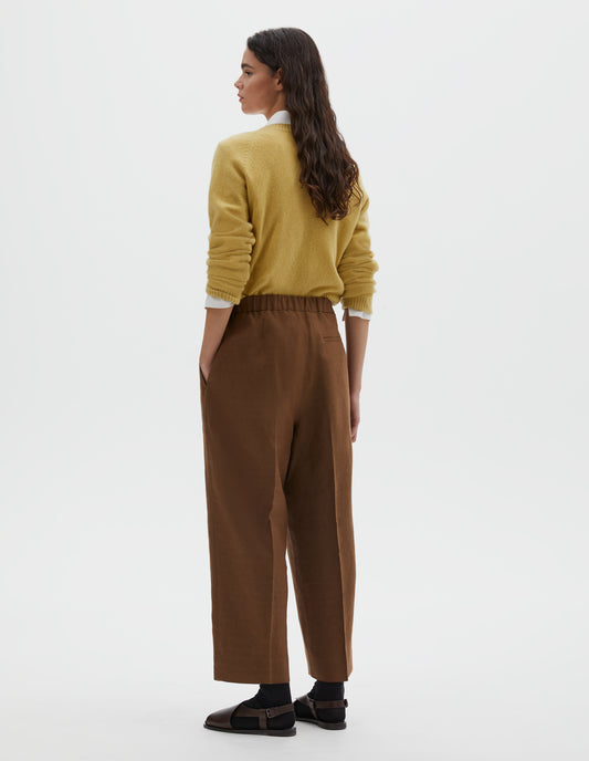 Full body image, back view of model wearing  brown linen trousers, with half elasticated waistband and belt back pocket.  Styled with  yellow long sleeve jumper, over white cotton shirt, with black ankle socks and brown leather sandals.