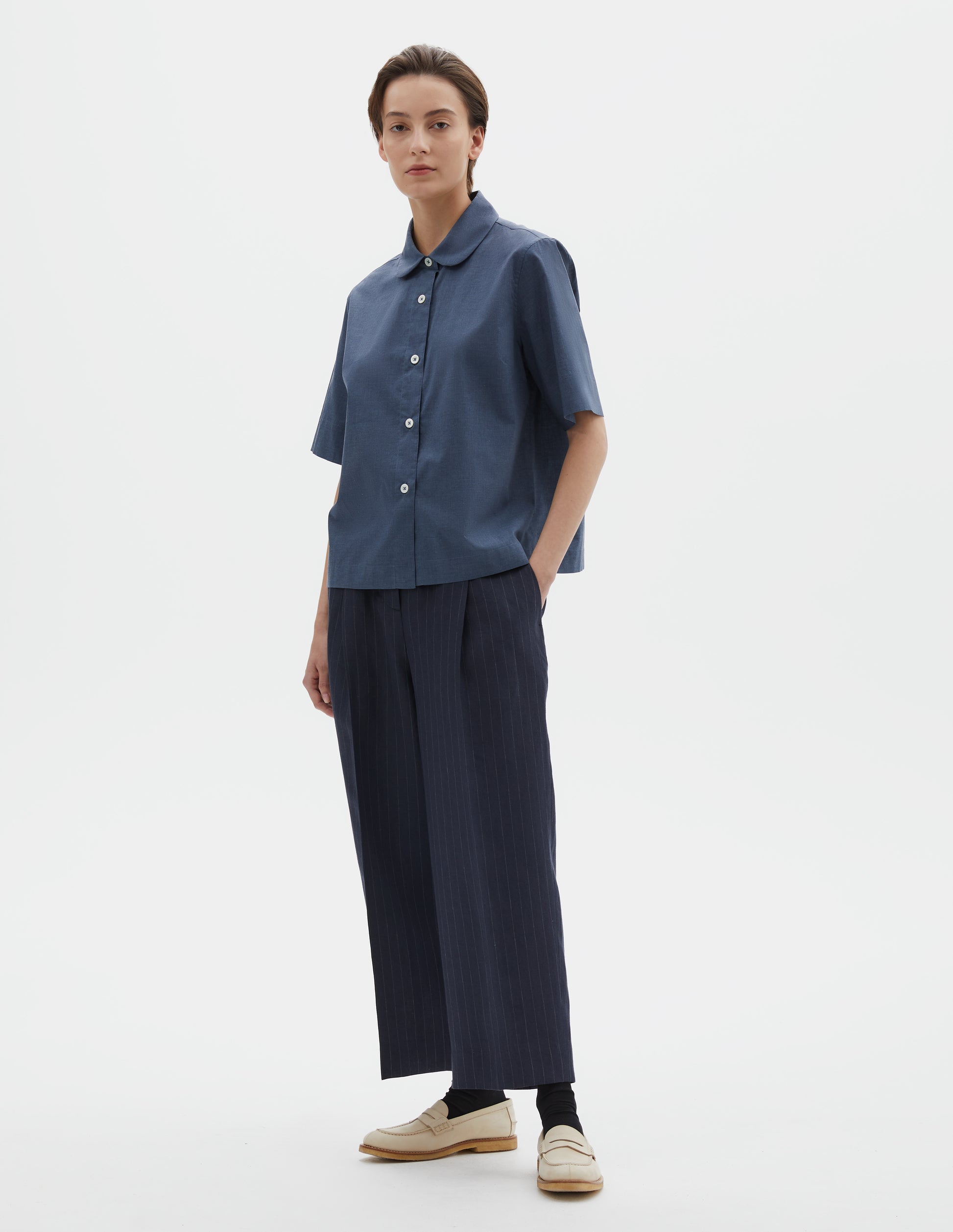 Full body image, front view of model wearing light blue cotton short sleeve shirt, with rounded collar, mother of pearl buttons and side split hem. Styled with navy blue and grey pinstripe trousers, black socks and beige suede loafers.