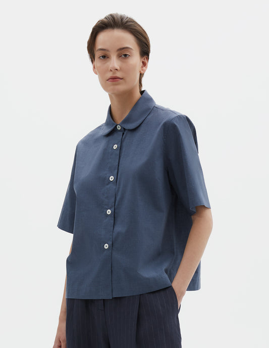 Cropped body image, front view of model wearing light blue cotton short sleeve shirt, with rounded collar, mother of pearl buttons and side split hem. Styled with navy blue and grey pinstripe trousers.