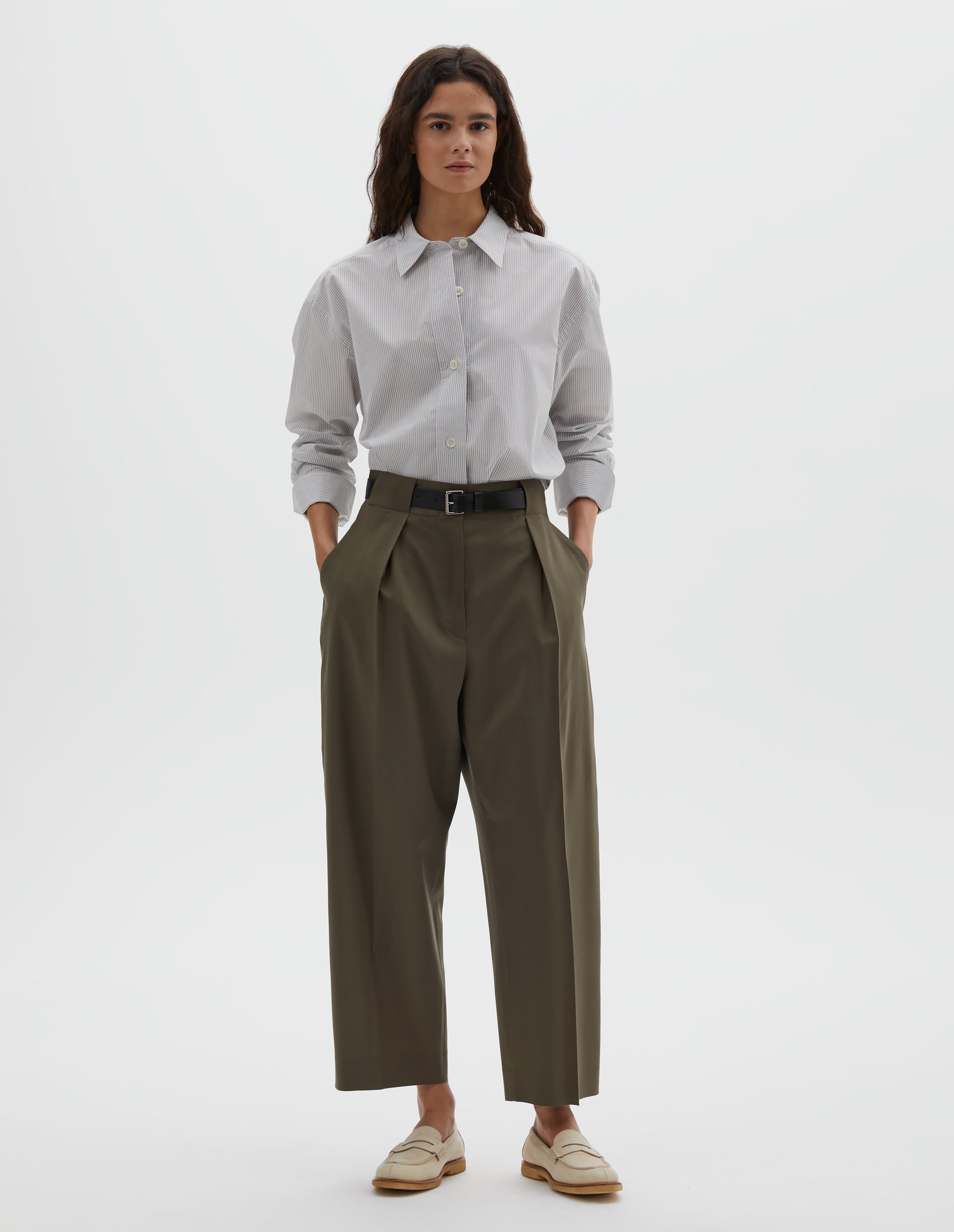 Full body image, front view of model wearing brown wool trousers, single pleats and grown-on waistband with large belt loops and horn button. Styled with grey shirt , dark brown belt and beige shoes.