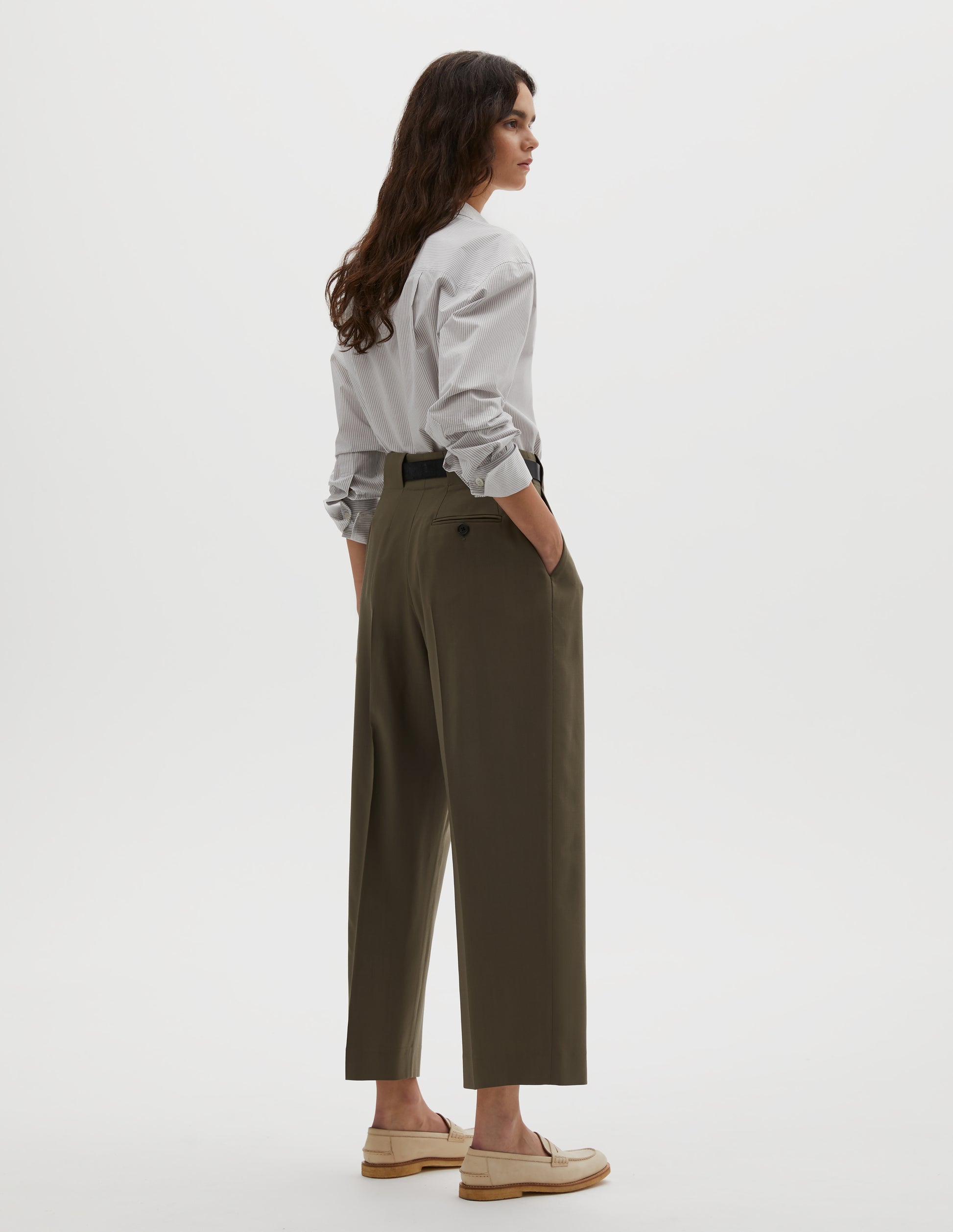 Full body image, back view of model wearing brown wool trousers, grown-on waistband with large belt loops and a button-through jet back pocket and horn button. Styled with grey shirt , dark brown belt and beige shoes.