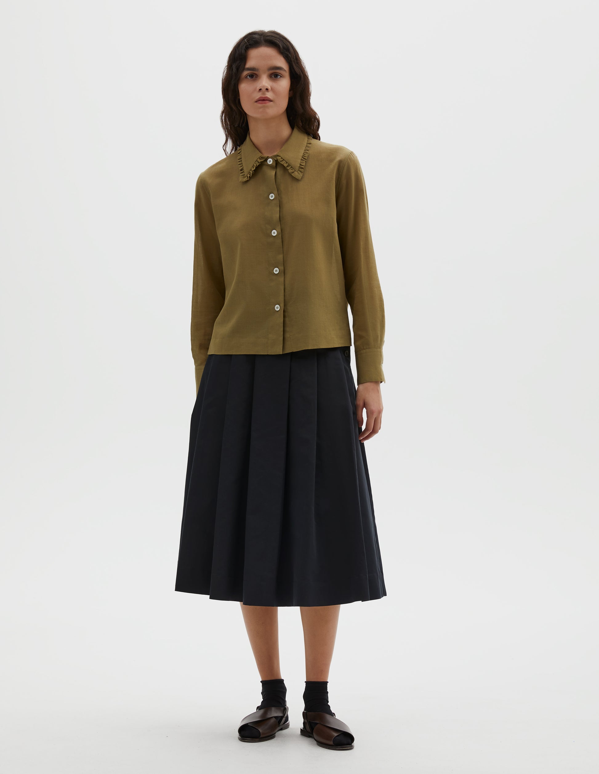 Full body image, front view of model wearing green cotton cashmere frilled collar long sleeve shirt, with mother of pearl button closure. Styled with pleated navy blue skirt, black ankle socks and brown leather sandals.