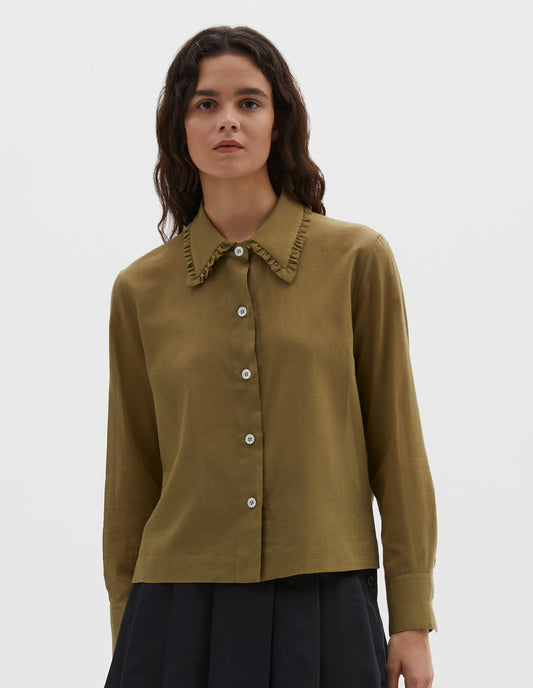 Cropped body image, front view of model wearing green cotton cashmere frilled collar long sleeve shirt, with mother of pearl button closure. Styled with pleated navy blue skirt.