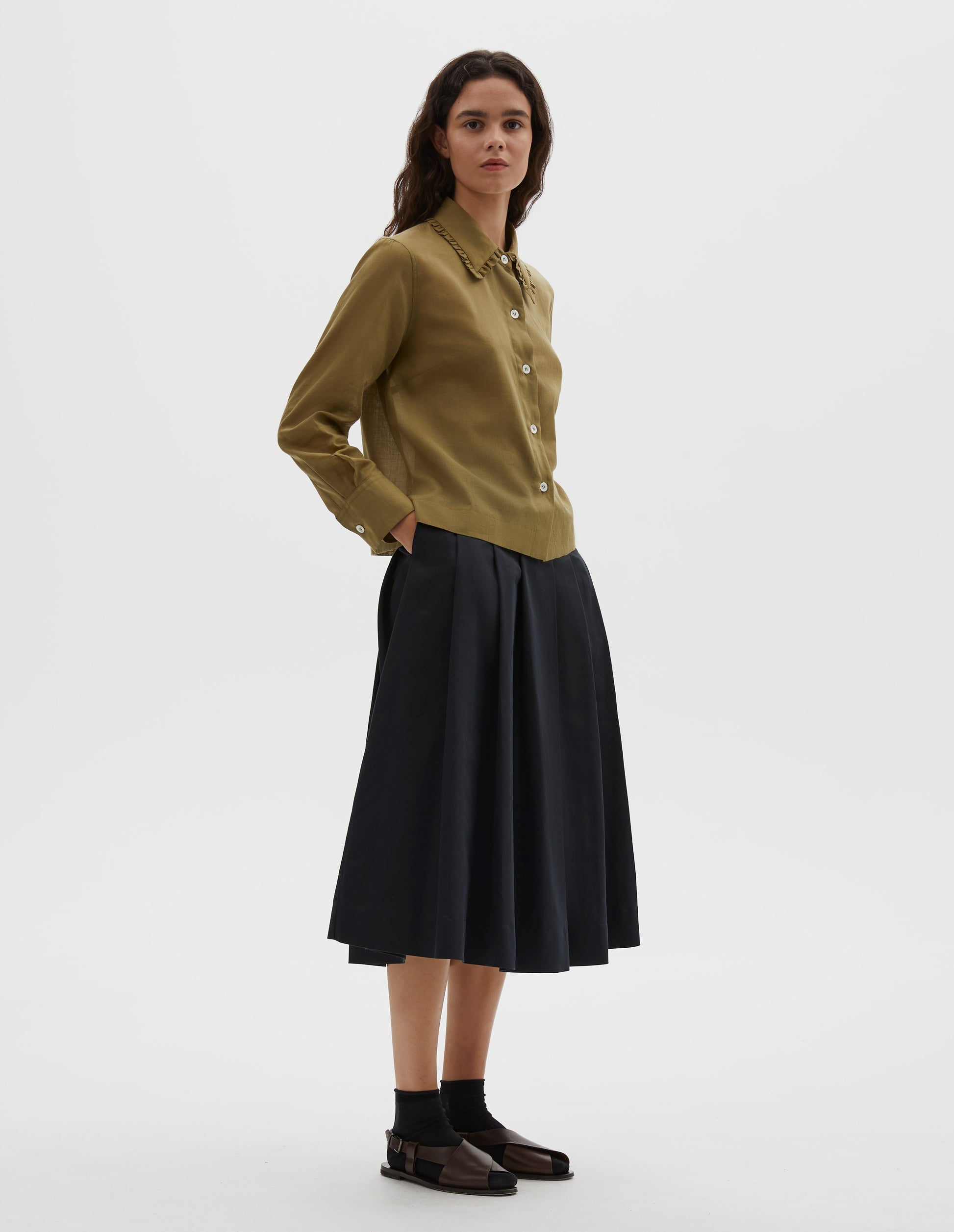 Full body image, front view of model wearing black relaxed cut below knee skirt, minimal waistband and side button fastening, stitched down pleats, and concealed side seam pockets. Styled with brownish green shirt, black socks and brown leather sandals.