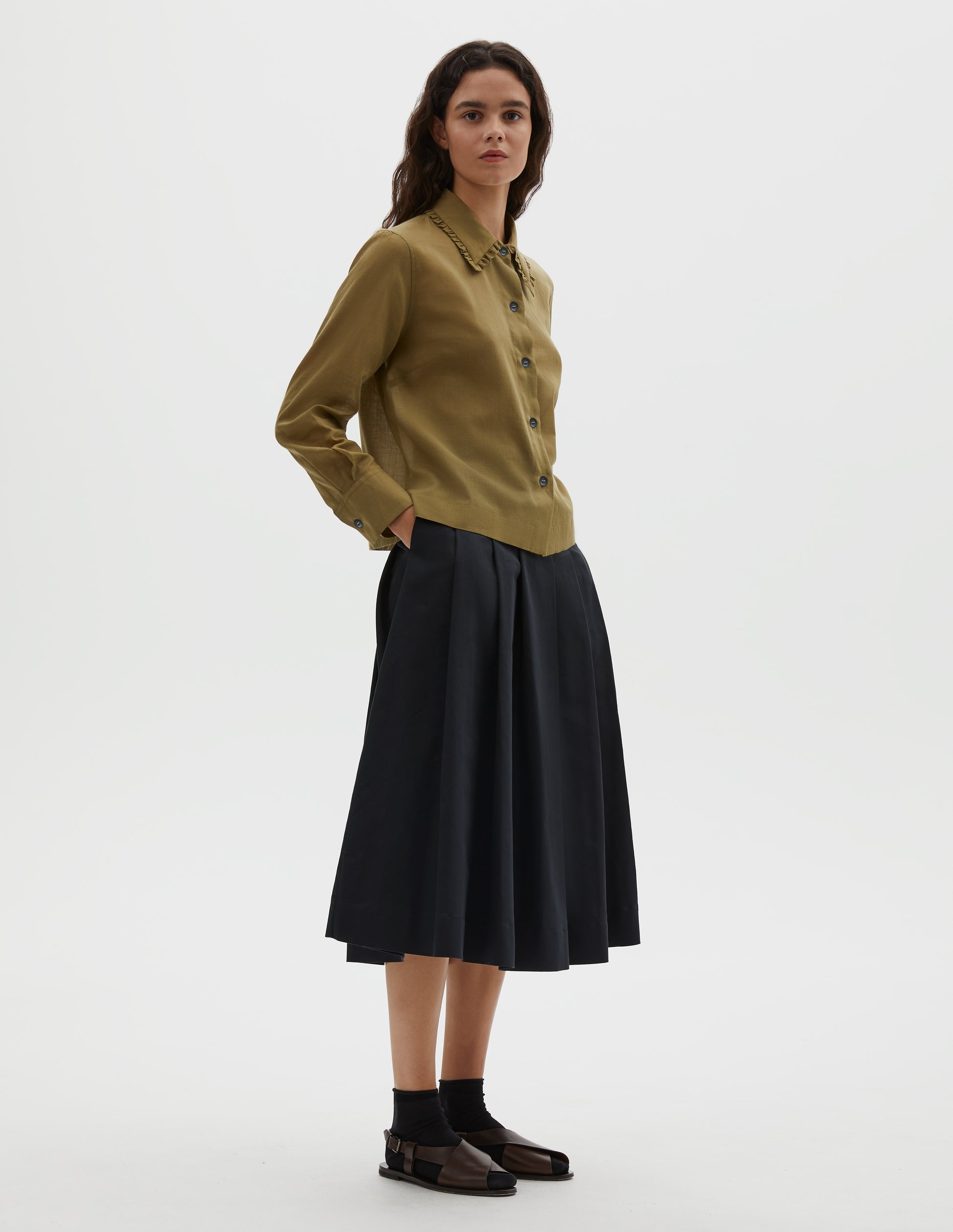 Full body image, side view of model wearing green cotton cashmere frilled collar long sleeve shirt, with horn button closure. Styled with pleated black skirt, black ankle socks and brown leather sandals.