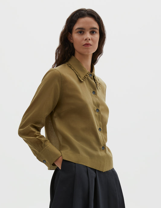 Cropped body image, side view of model wearing green cotton cashmere frilled collar long sleeve shirt, with horn button closure. Styled with pleated black skirt.