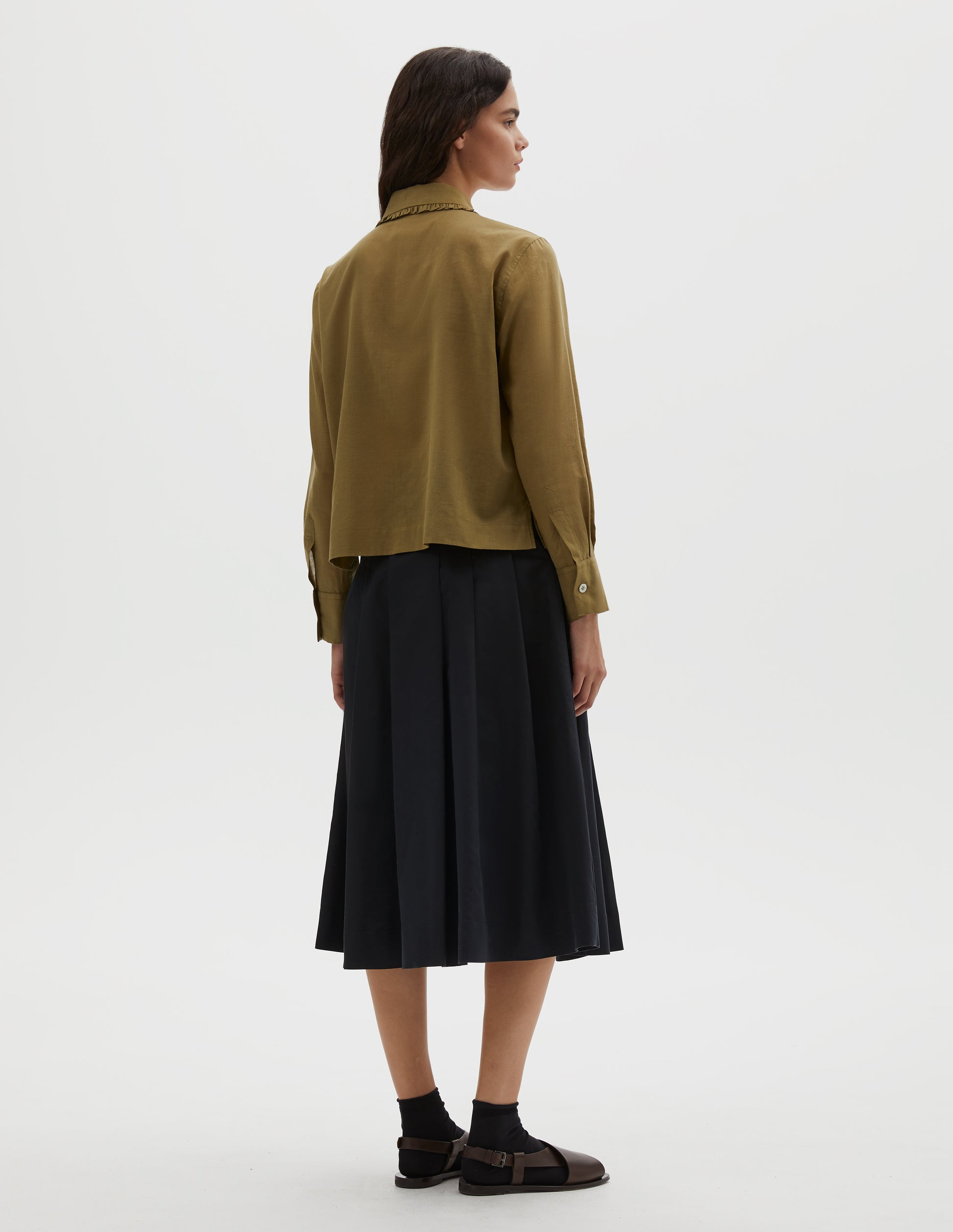 Full body image, back view of model wearing green cotton cashmere frilled collar long sleeve shirt, with mother of pearl button cuffs. Styled with navy blue pleated skirt, black ankle socks and brown leather sandals.