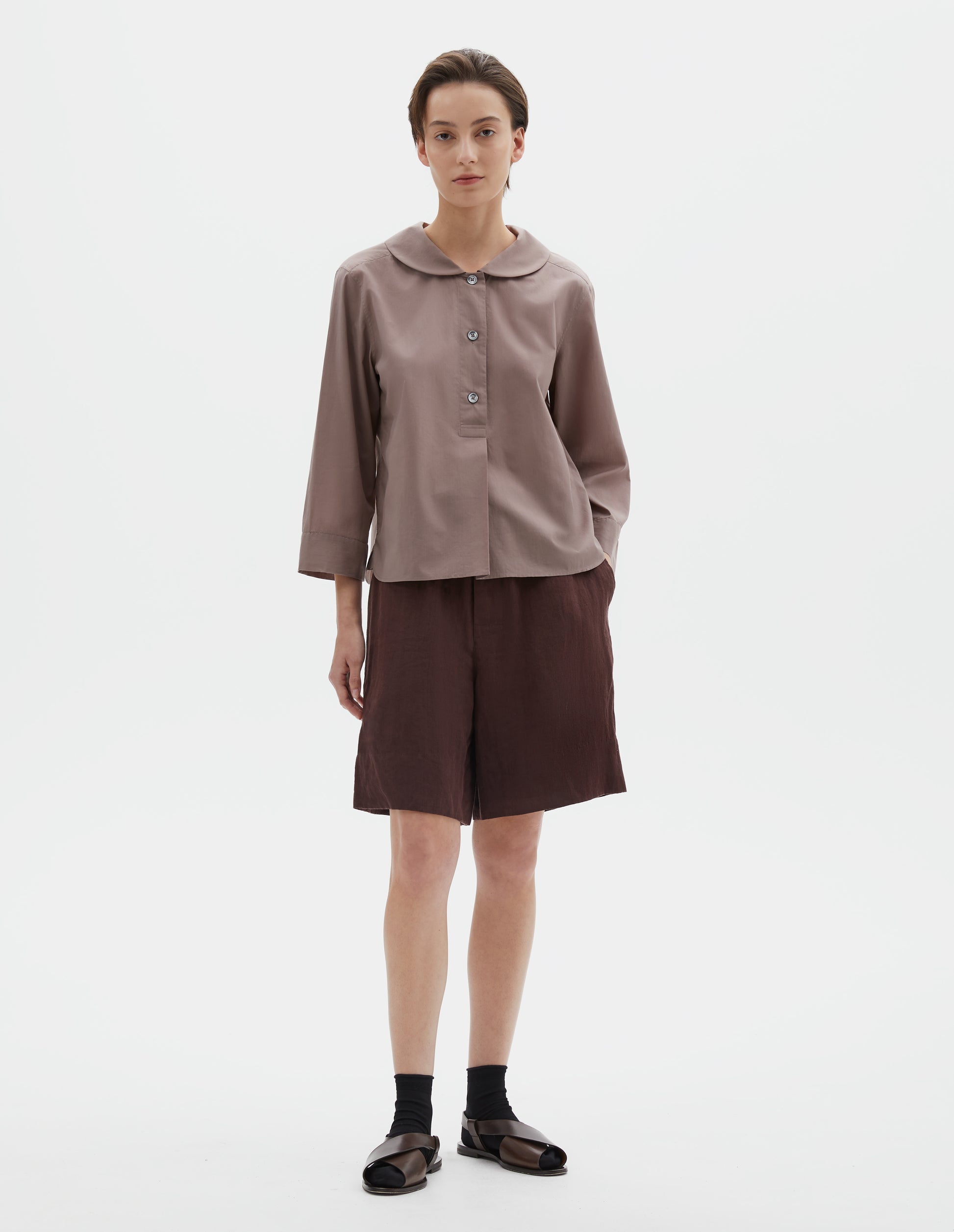 Full body image, front view of model wearing pinkish brown pull-on shirt with rounded collar, stitch-through half placket and three quarter sleeves with a turned back cuff detail. Styled with brown shorts and black socks with dark brown leather sandals.