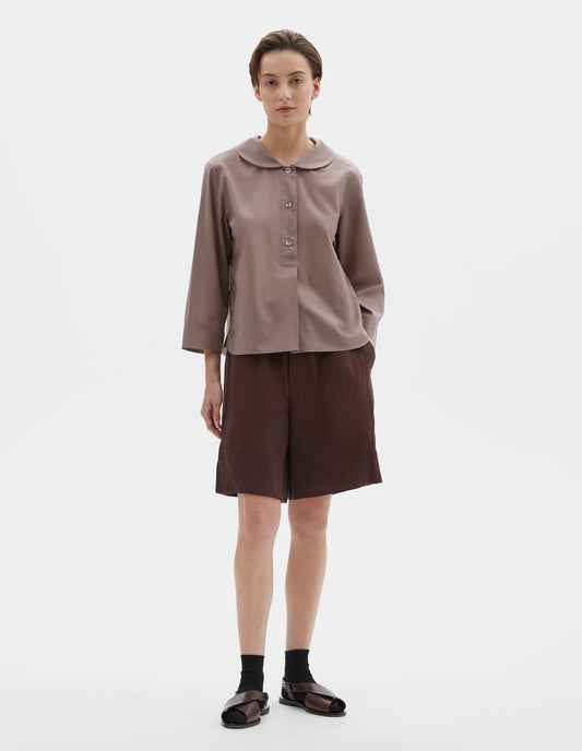 Full body image, front view of model wearing pinkish brown pull-on shirt with rounded collar, stitch-through half placket and three quarter sleeves with a turned back cuff detail. Styled with brown shorts and black socks with dark brown leather sandals.
